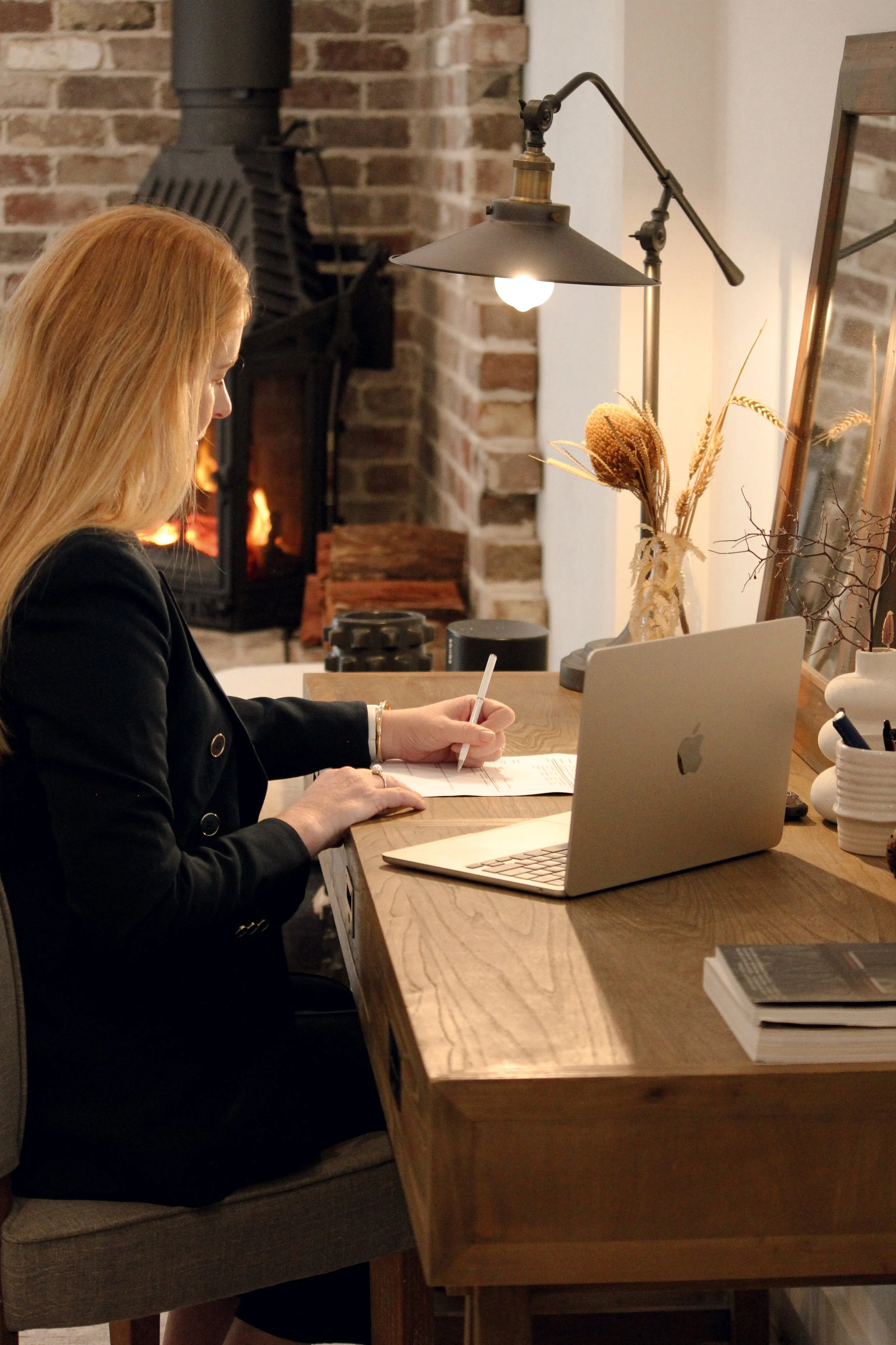 Woman working at a wooden desk with an open laptop, notebooks, and decorative dried flowers, in a cozy room with a brick fireplace. Louise Neilson - Neilson and Co Legal - The Hunter Valley, Maitland and Newcastle.