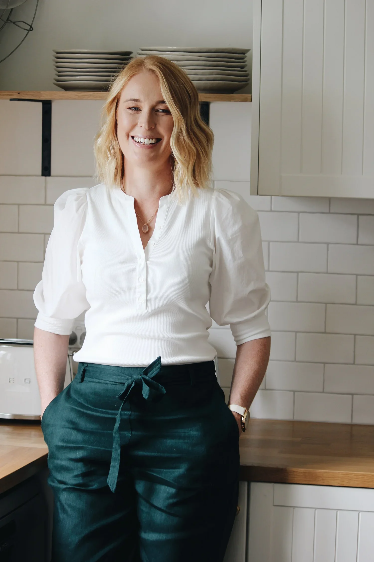 A woman with shoulder-length blonde hair smiling in a modern kitchen, wearing a white blouse and dark green pants. Louise Neilson - Neilson and Co Legal - The Hunter Valley, Maitland and Newcastle.
