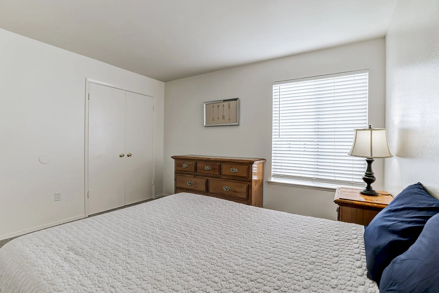 This image shows a brightly lit bedroom featuring a bed dressed in a textured white quilt and dark blue pillows. Traditional wooden furniture, including a dresser and a matching nightstand. 