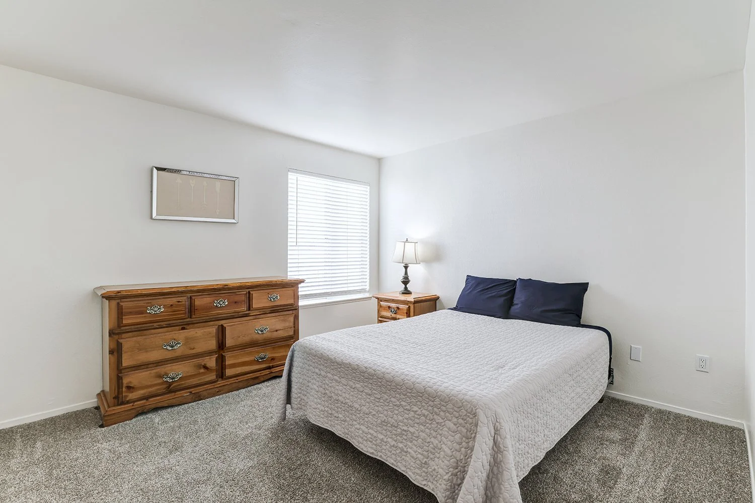 This image shows a brightly lit bedroom featuring a bed dressed in a textured white quilt and dark blue pillows. Traditional wooden furniture, including a dresser and a matching nightstand. 