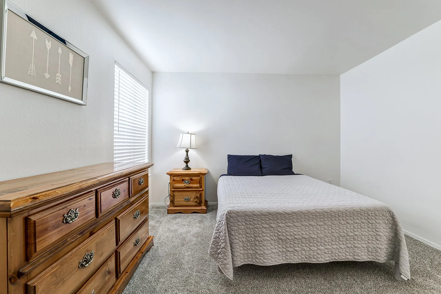 This image shows a brightly lit bedroom featuring a bed dressed in a textured white quilt and dark blue pillows. Traditional wooden furniture, including a dresser and a matching nightstand. 