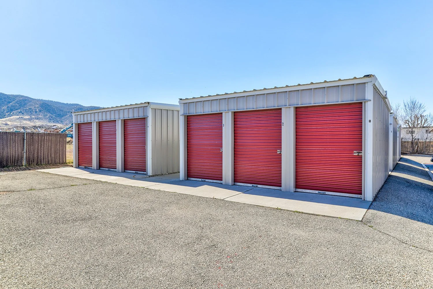 This image shows rows of outdoor self-storage units with metallic siding and bold red roll-up doors facing a driveway.