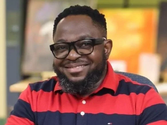 A smiling man with glasses and a beard, wearing a red and navy striped polo shirt, seated in an indoor setting with a blurred background.