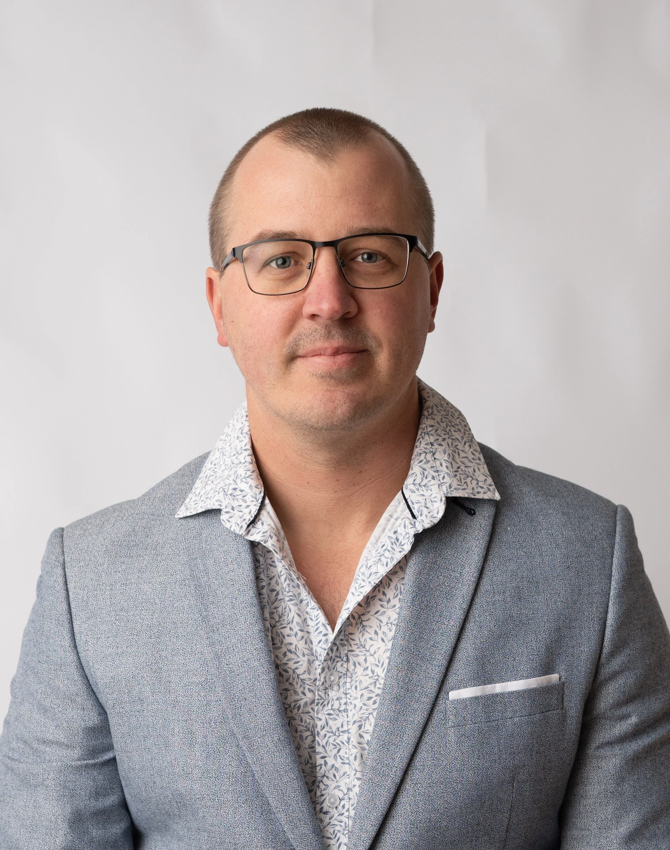 A man with glasses wearing a patterned shirt and a gray blazer against a plain light-colored background.