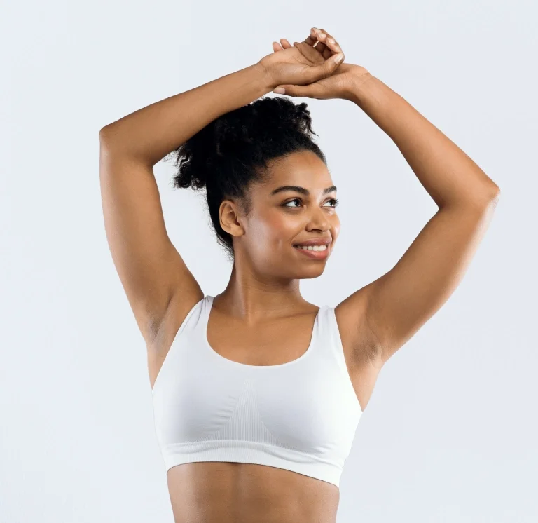 Young woman stretching with arms above head, smiling, wearing a white sports bra, against a plain white background.