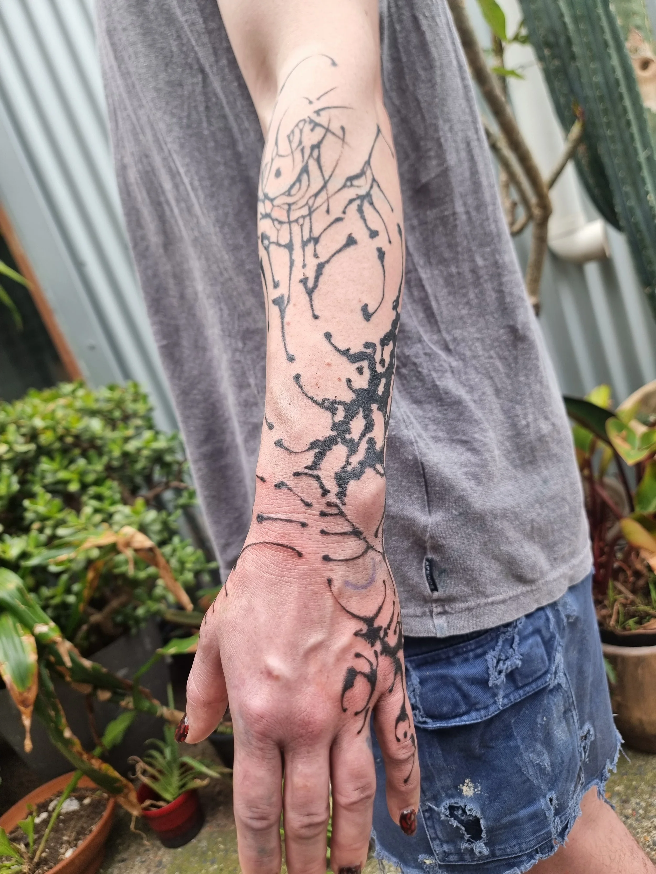 A person's arm with a large black tattoo of a dragon or mythical creature. The person is wearing a gray shirt and ripped denim shorts, with a garden background.