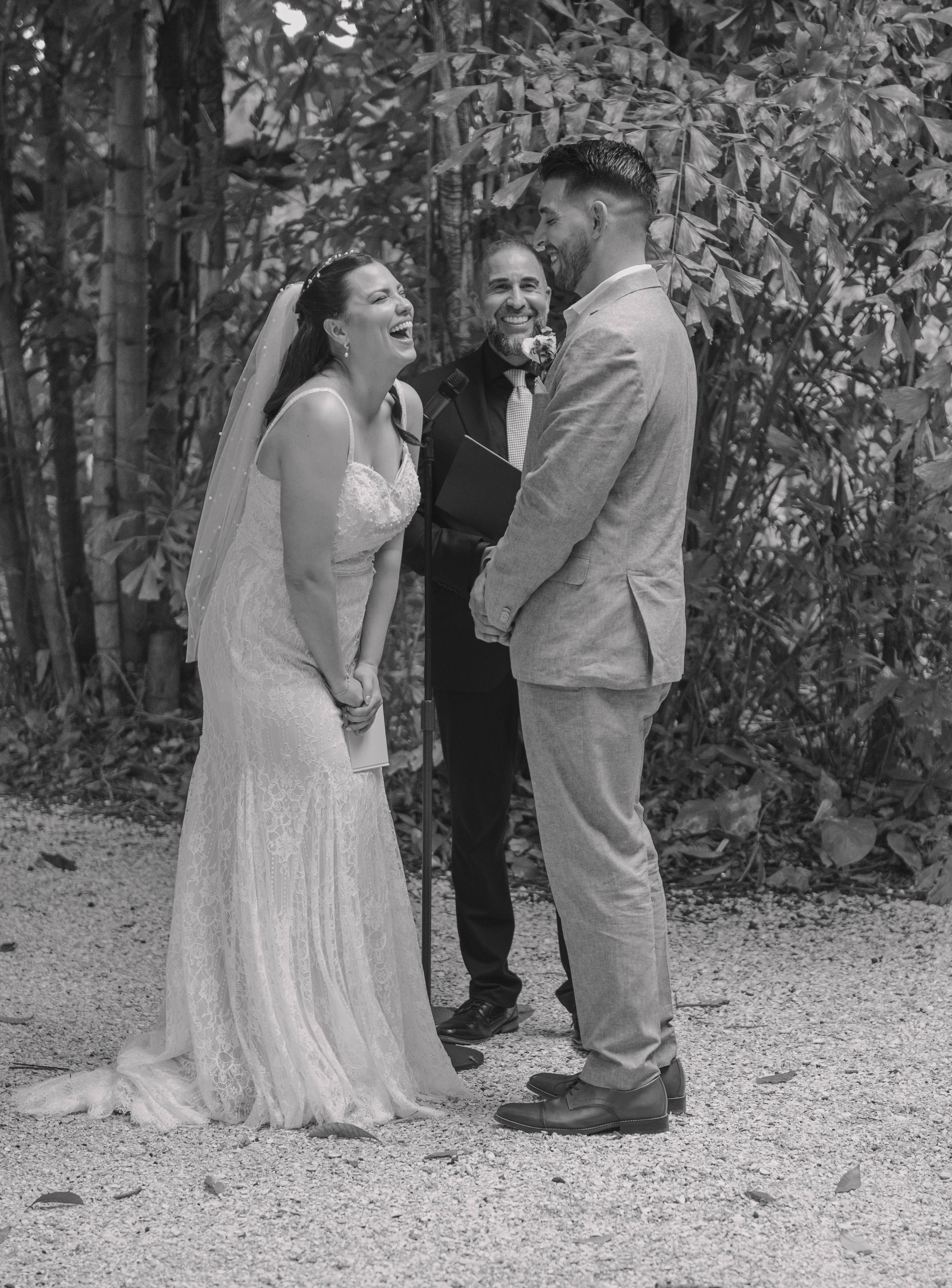 A bride and groom laughing during an outdoor wedding ceremony with an officiant standing between them.