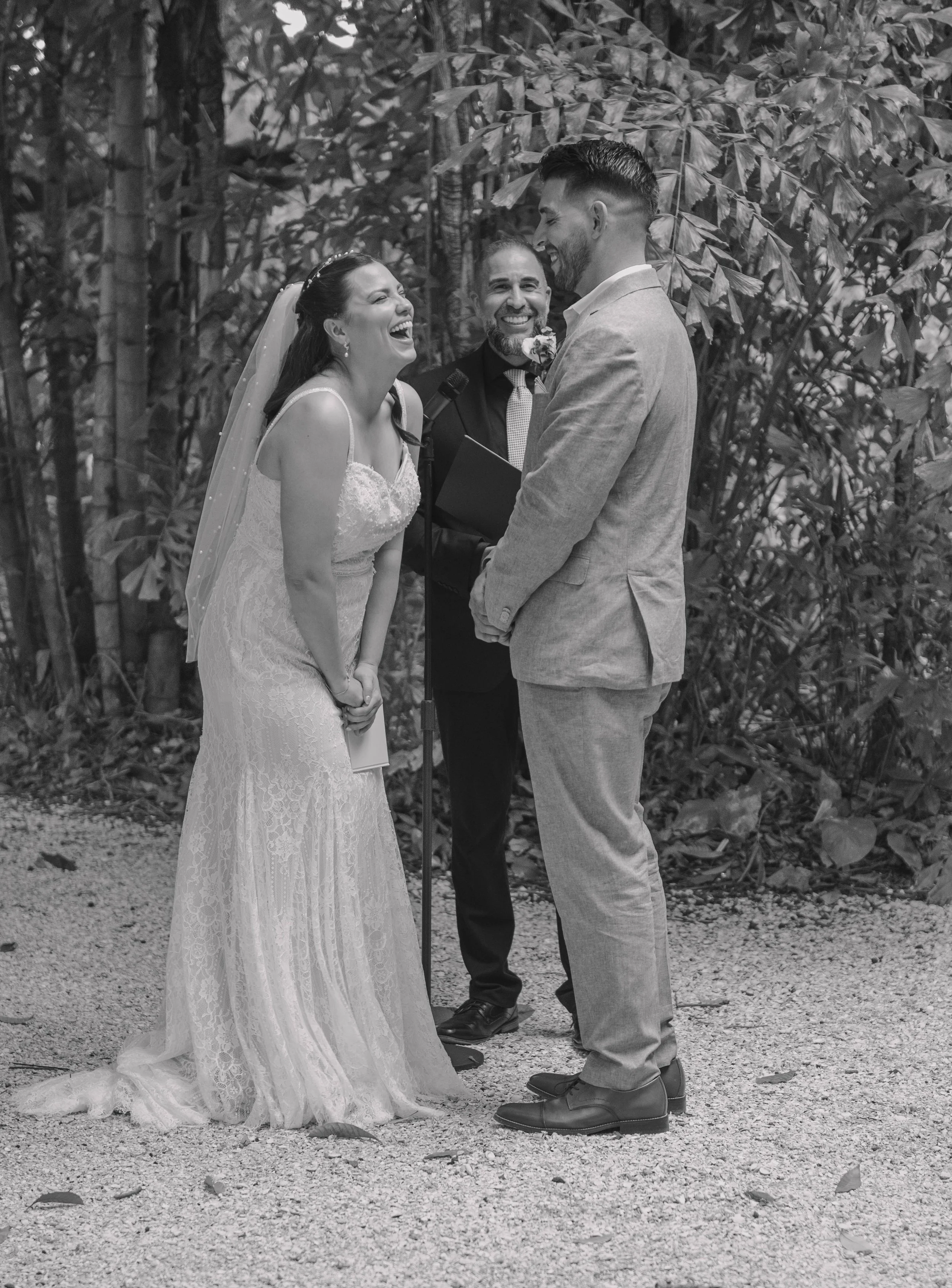 A wedding ceremony outdoors with a bride and groom smiling at each other, an officiant smiling in the background, surrounded by trees.