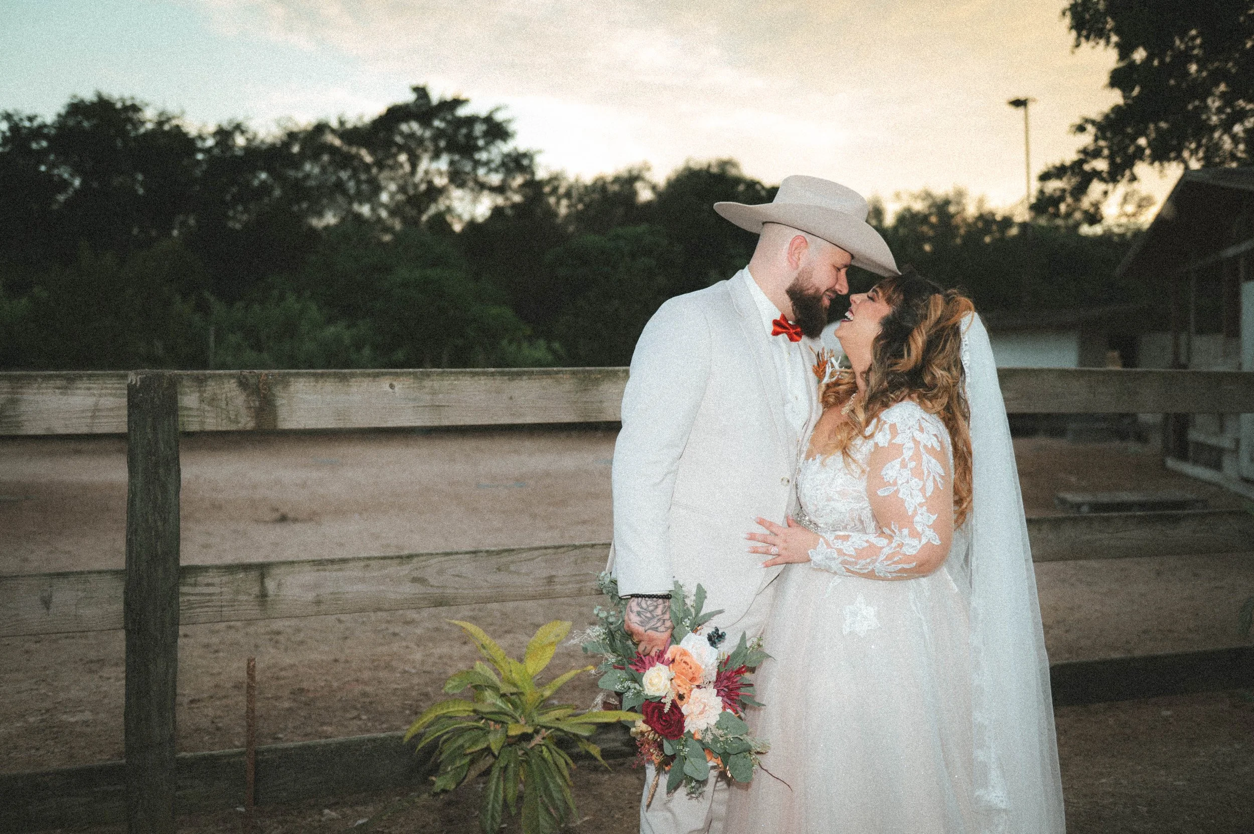 A bride and groom embrace outdoors at sunset, smiling at each other, with the bride holding a bouquet of flowers and the groom wearing a white suit and cowboy hat.