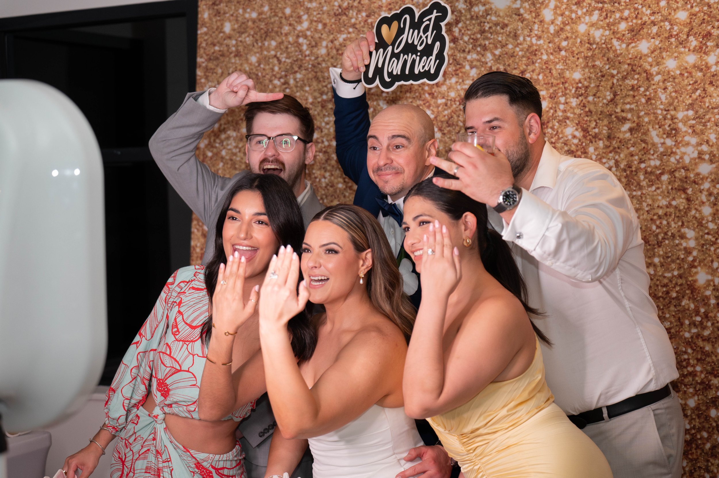 Group of people posing for a photo at a wedding, with "Just Married" sign, smiling and showing rings.