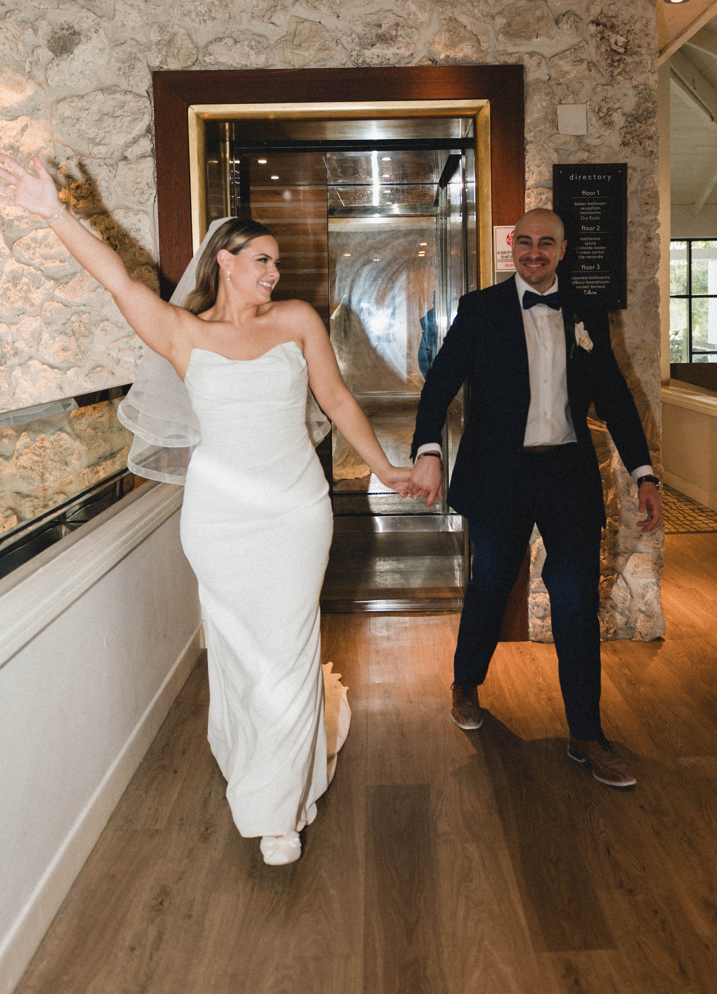 Bride and groom walking hand in hand, bride wearing a white dress and groom in a dark suit, celebrating indoors.