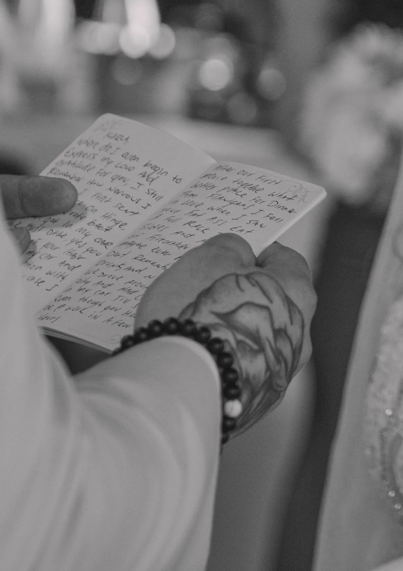 A hand with a tattoo on the wrist holding a small open notebook with handwritten notes inside. The background is out of focus with soft bokeh lights.