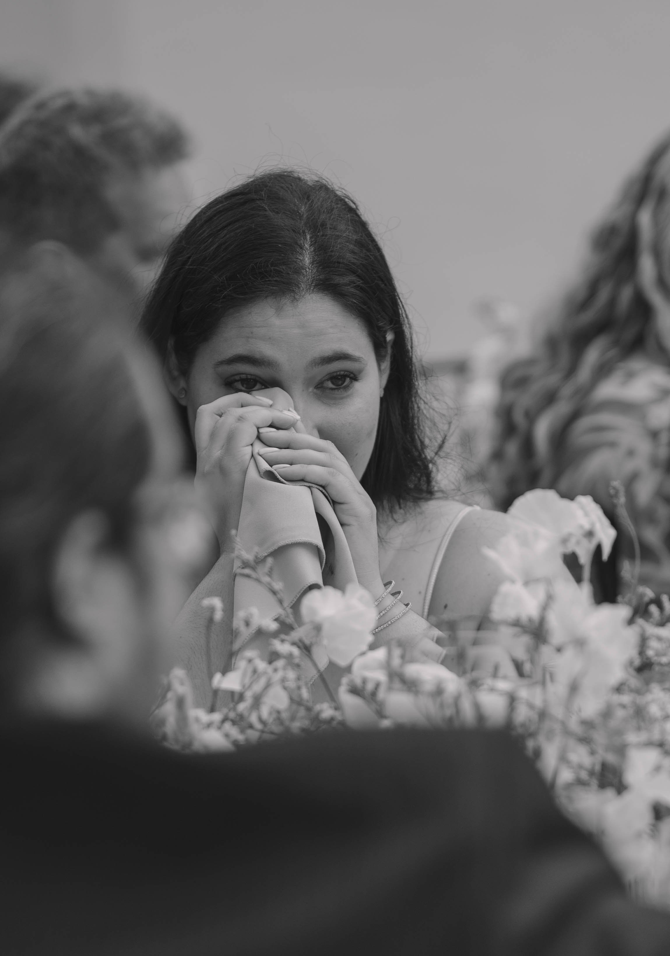 Black and white photo of a woman at a table wiping her eyes with a napkin, surrounded by flowers, with other people blurred in the background.