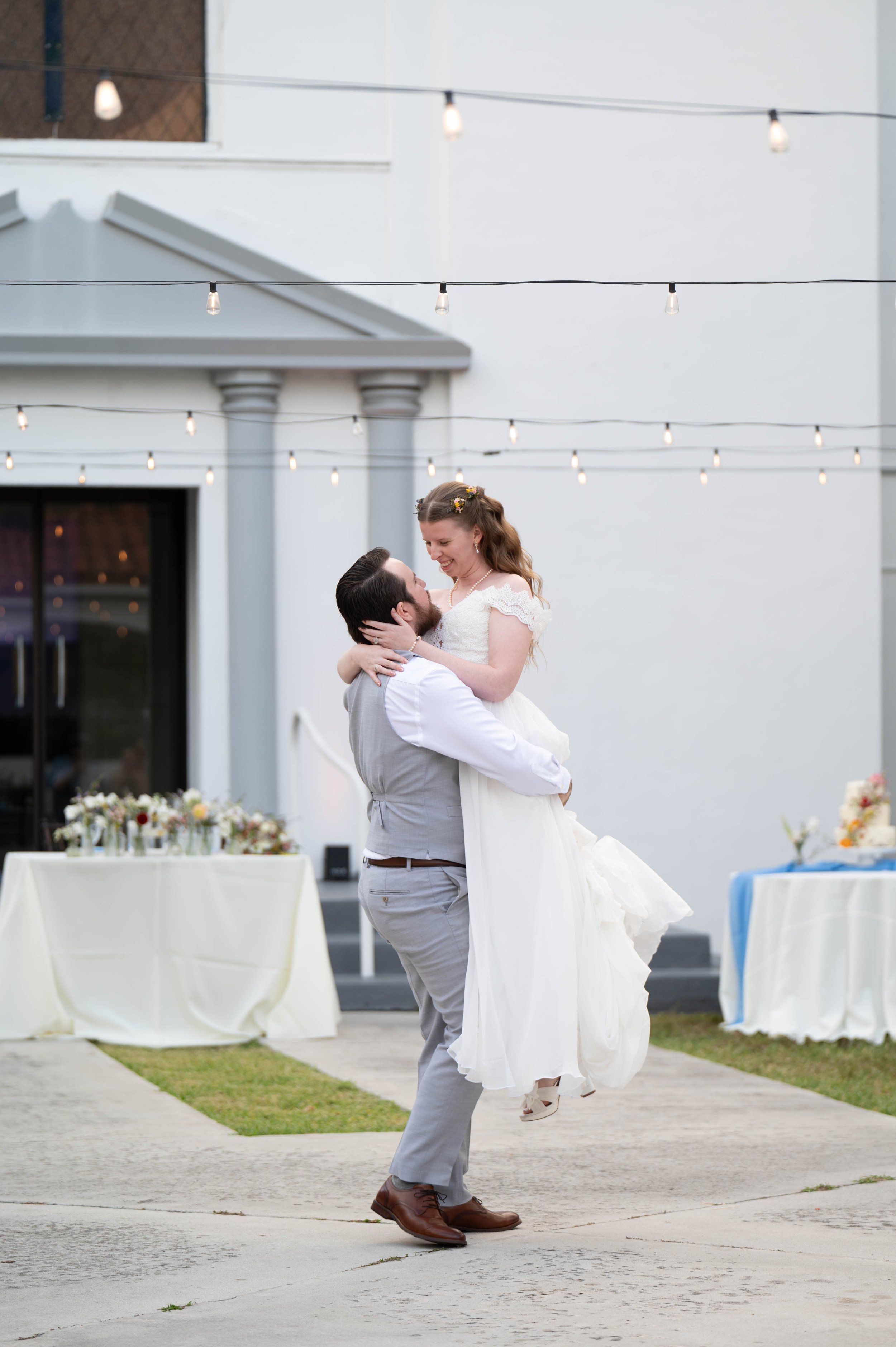 Bride and groom outdoors at a wedding, with groom lifting bride, surrounded by tables and string lights.