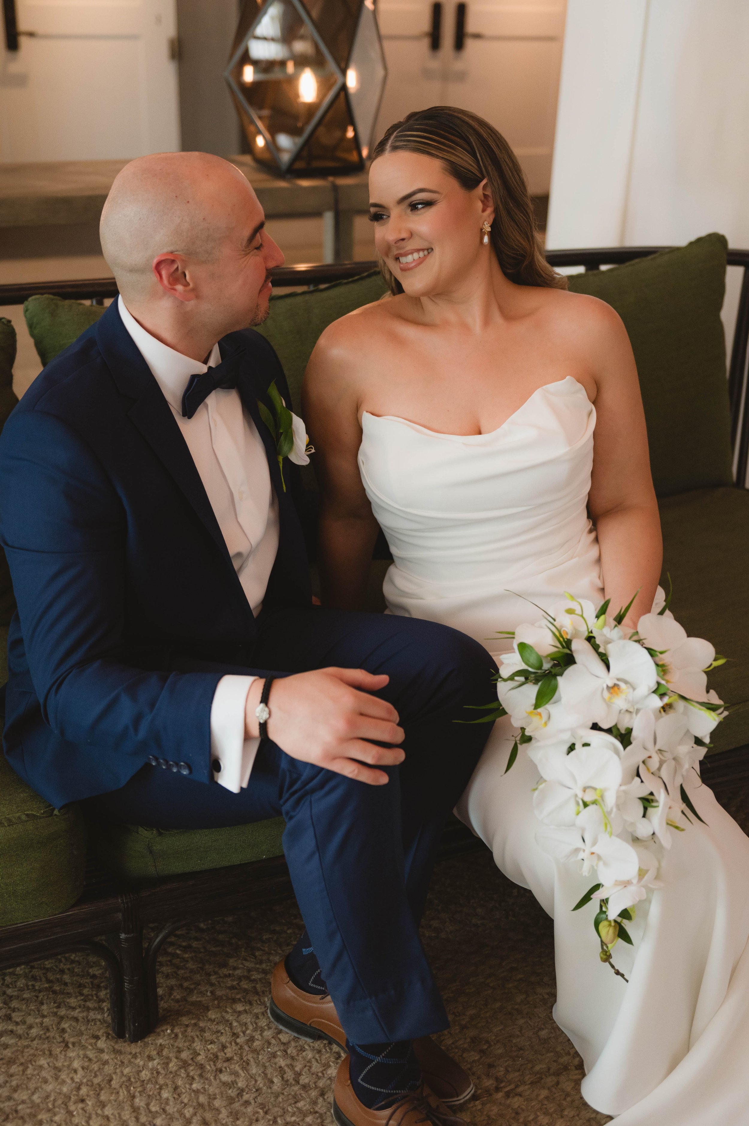A bride and groom sitting on a green couch, with the groom wearing a navy suit and the bride in a white dress holding a bouquet of white flowers. They are smiling at each other.
