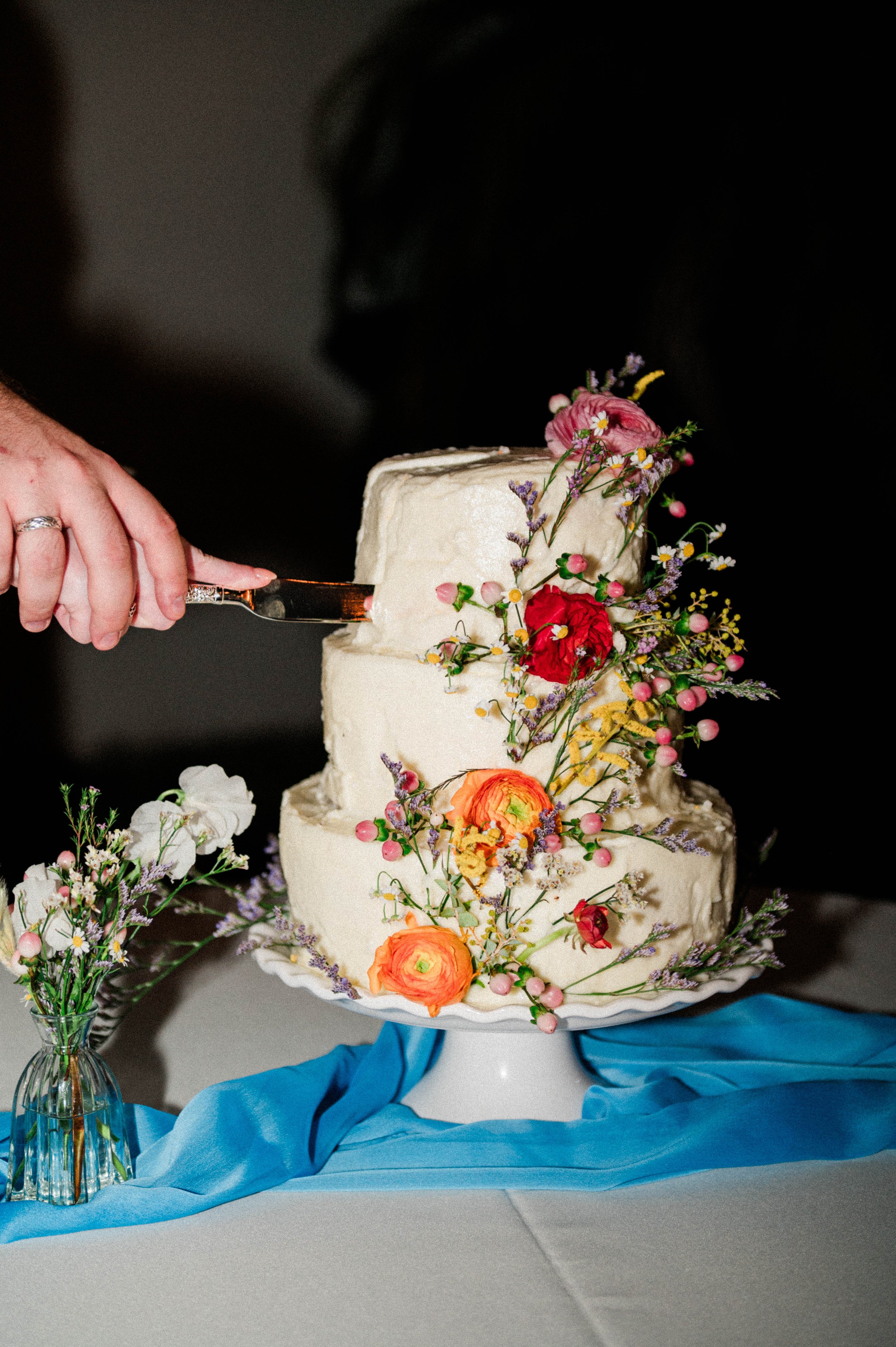 Two-tiered white cake with colorful flowers, being sliced with a knife, on a table with blue cloth and small bouquet.