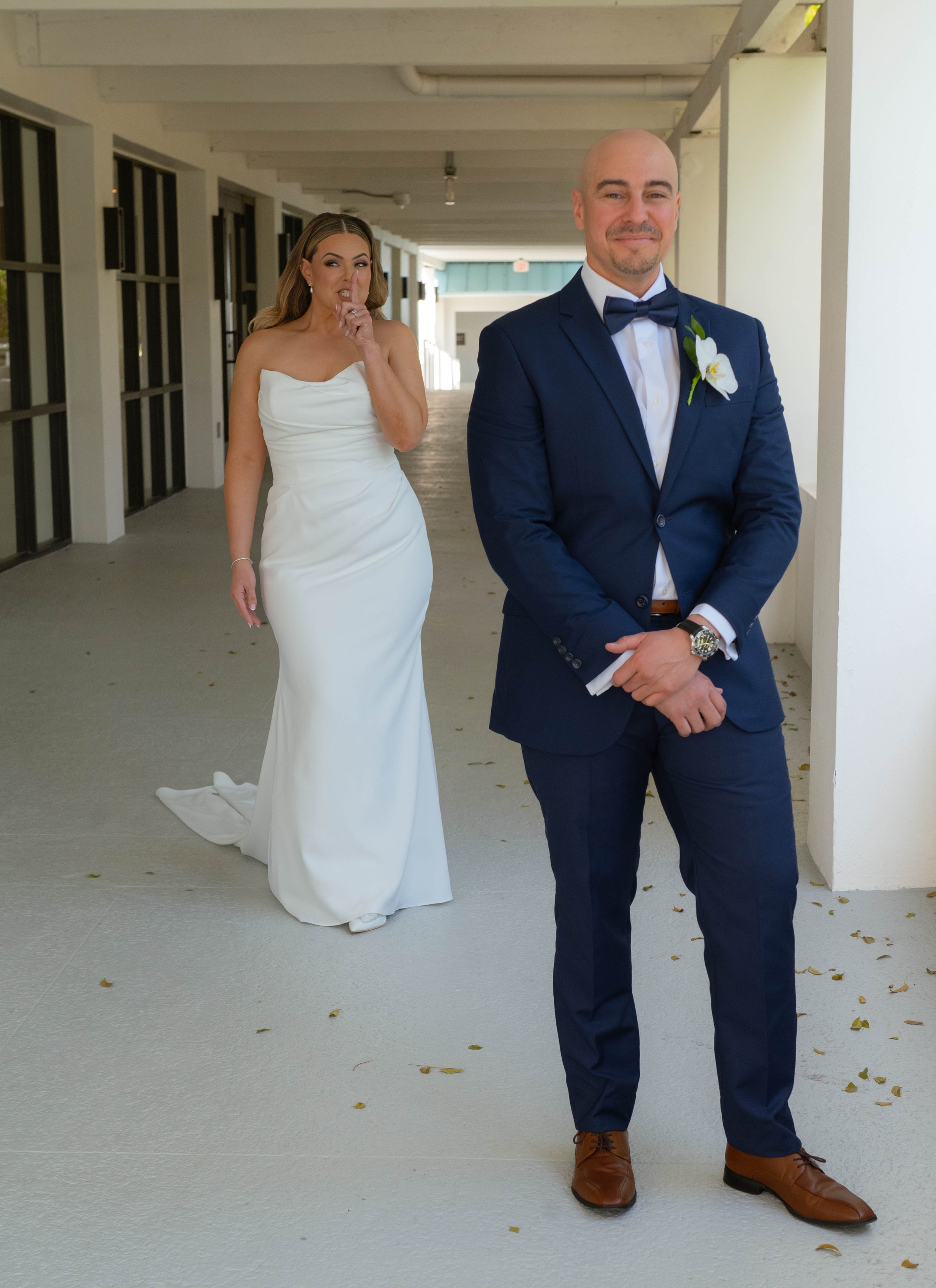 Bride playfully approaching groom for a first look, outdoor setting, bride in a white gown, groom in a blue suit with brown shoes.