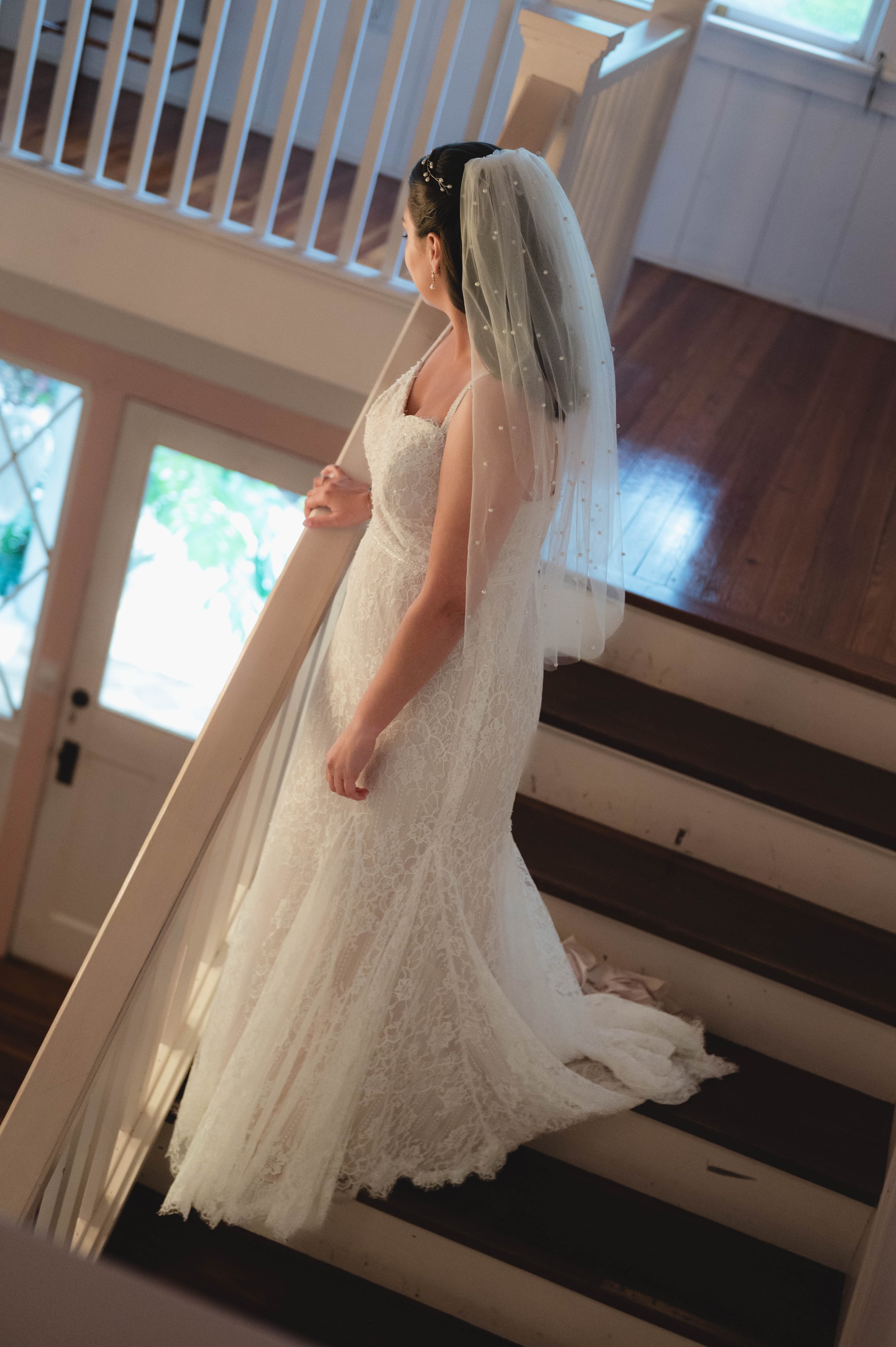 Bride in white lace wedding dress and veil standing on staircase indoors.