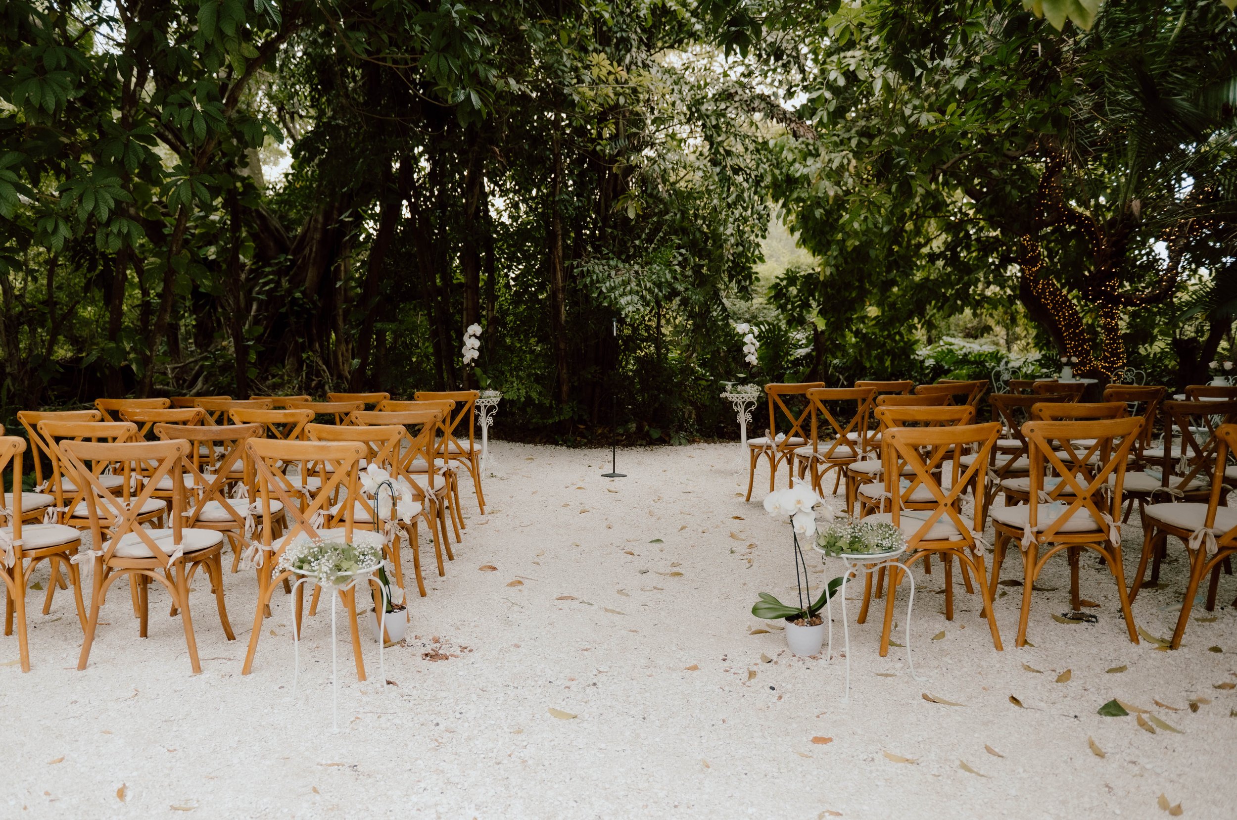 Outdoor wedding ceremony setup with wooden chairs and floral decorations in a garden setting under trees.