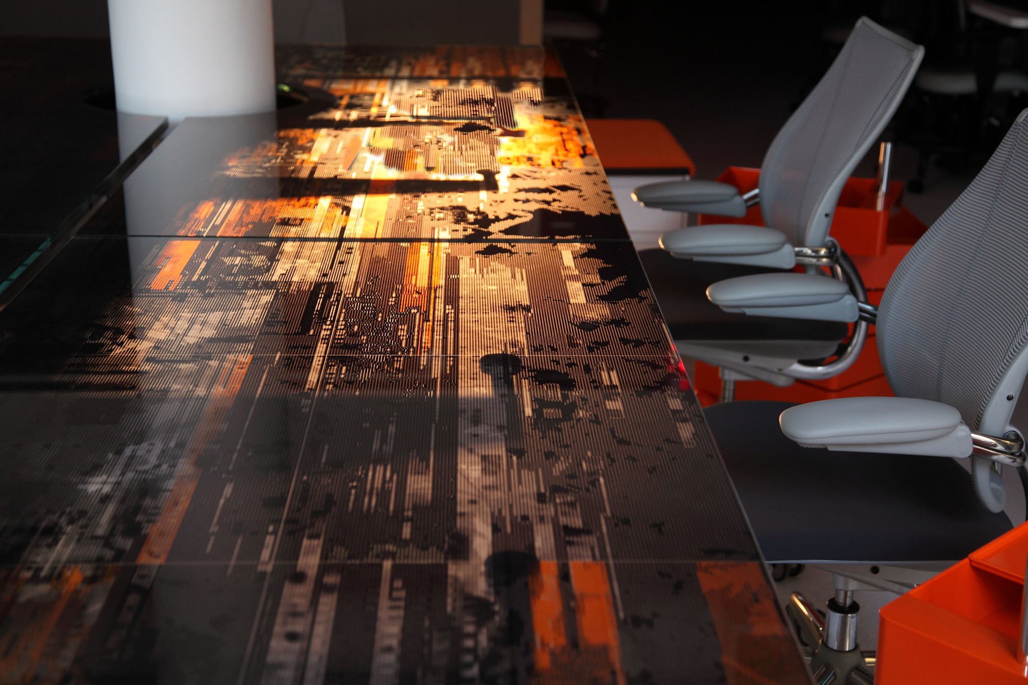 A steel conference table printed with a photograph ingrained sits in a conference room with two grey office chairs by it's side. The back of the table is lit with a warm LED light.