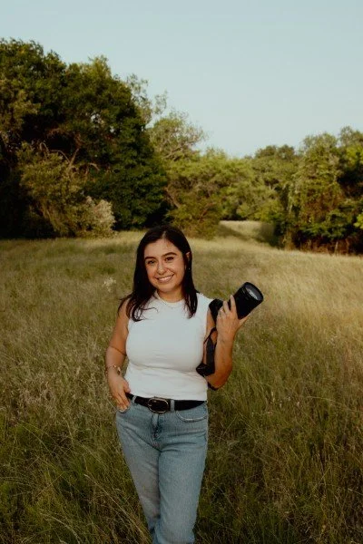 A young woman with dark hair wearing a white sleeveless top and blue jeans, holding a camera and smiling in a grassy field with trees in the background.