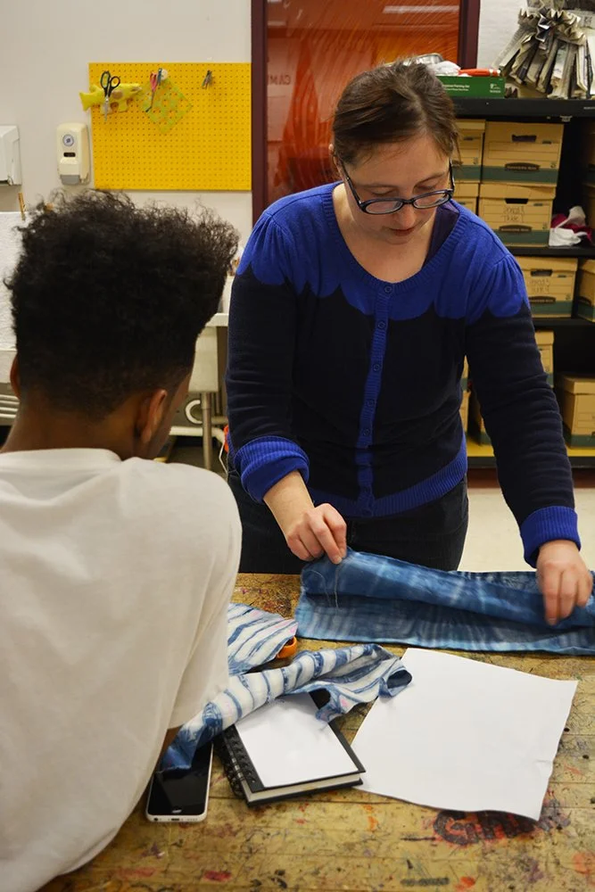 Teaching Artist Amber Coppings, in a blue sweater, demonstrates a shibori folding fabric technique to a student.