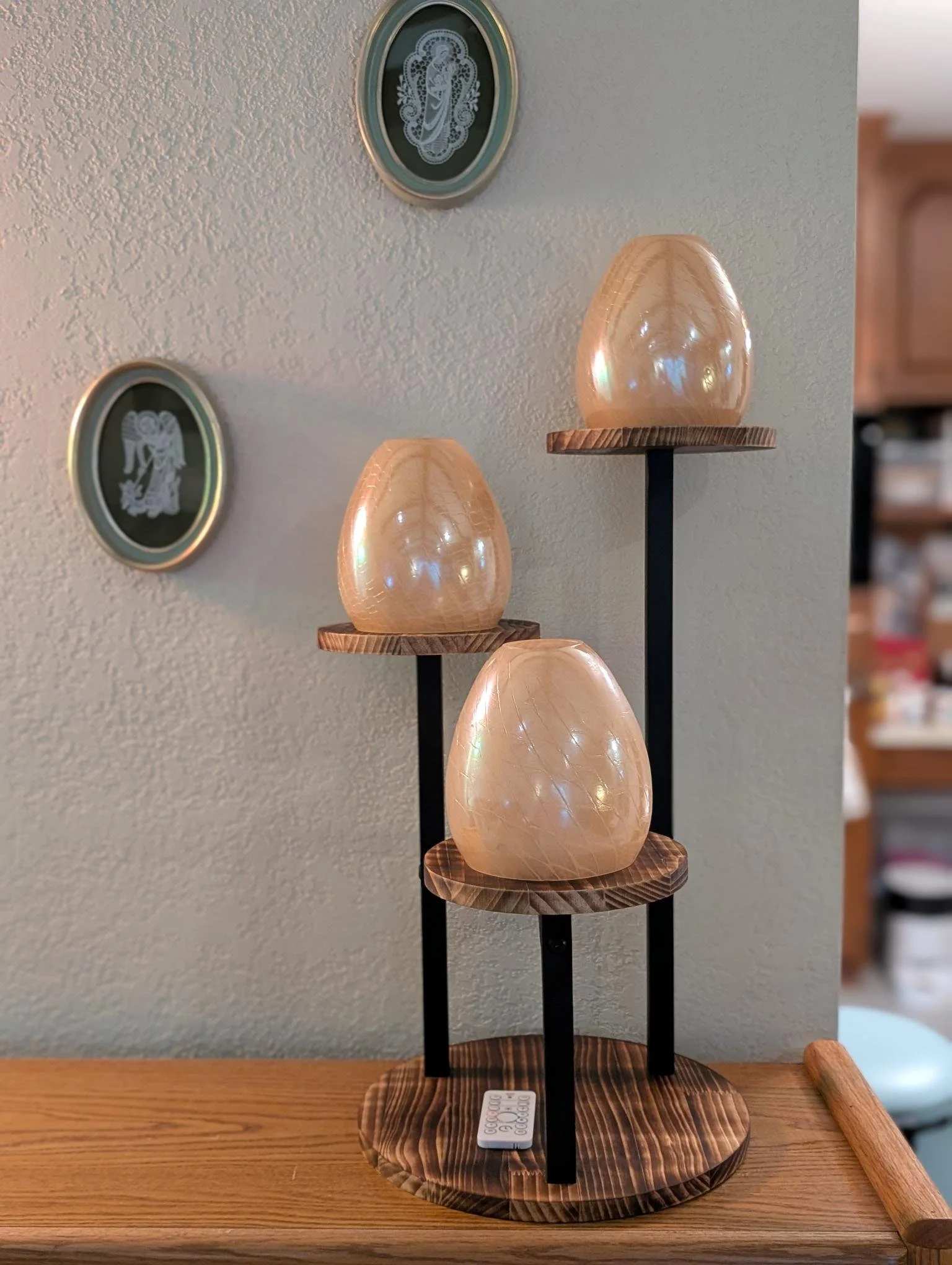 photo of a three tier plant stand with oyster colored glass cones. One cone is on each round wood platform. Platforms are supported by black iron. A white remote sits on the round base of the plant stand. On the wall behind are two framed lace pieces
