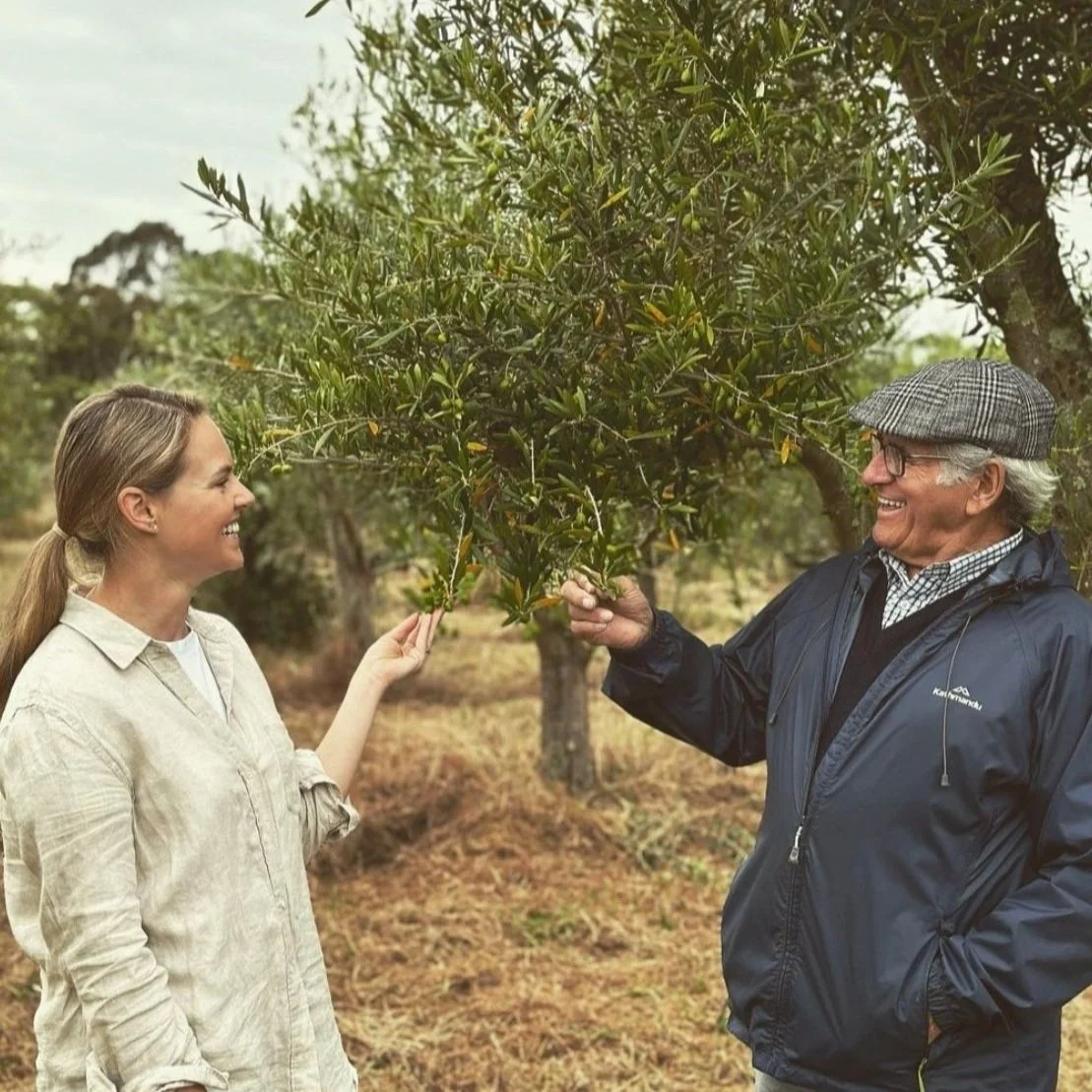 A woman and an older man are smiling and talking while standing outside near trees. The woman is holding a branch of an olive tree.