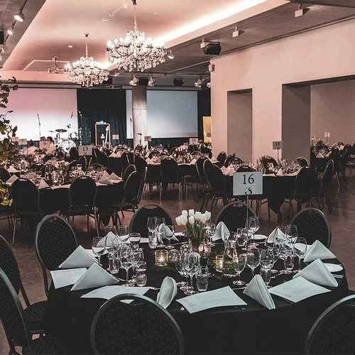 Bilde av et fint bord satt opp for et arrangement i en stor, elegant sal. Bordet har hvite duker, bestikk, glass, servietter og en blomsterdekorasjon. Flere andre bord er også satt opp i bakgrunnen, med tallkort, og det er store lysekroner hengende fra taket.
