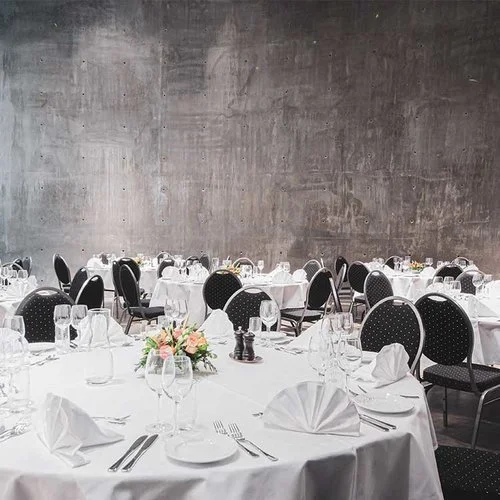 Festbord dekket med hvite duker, pyntet med blomstervase og bestikk, klare til et arrangement eller selskap i et rom med betongvegg.