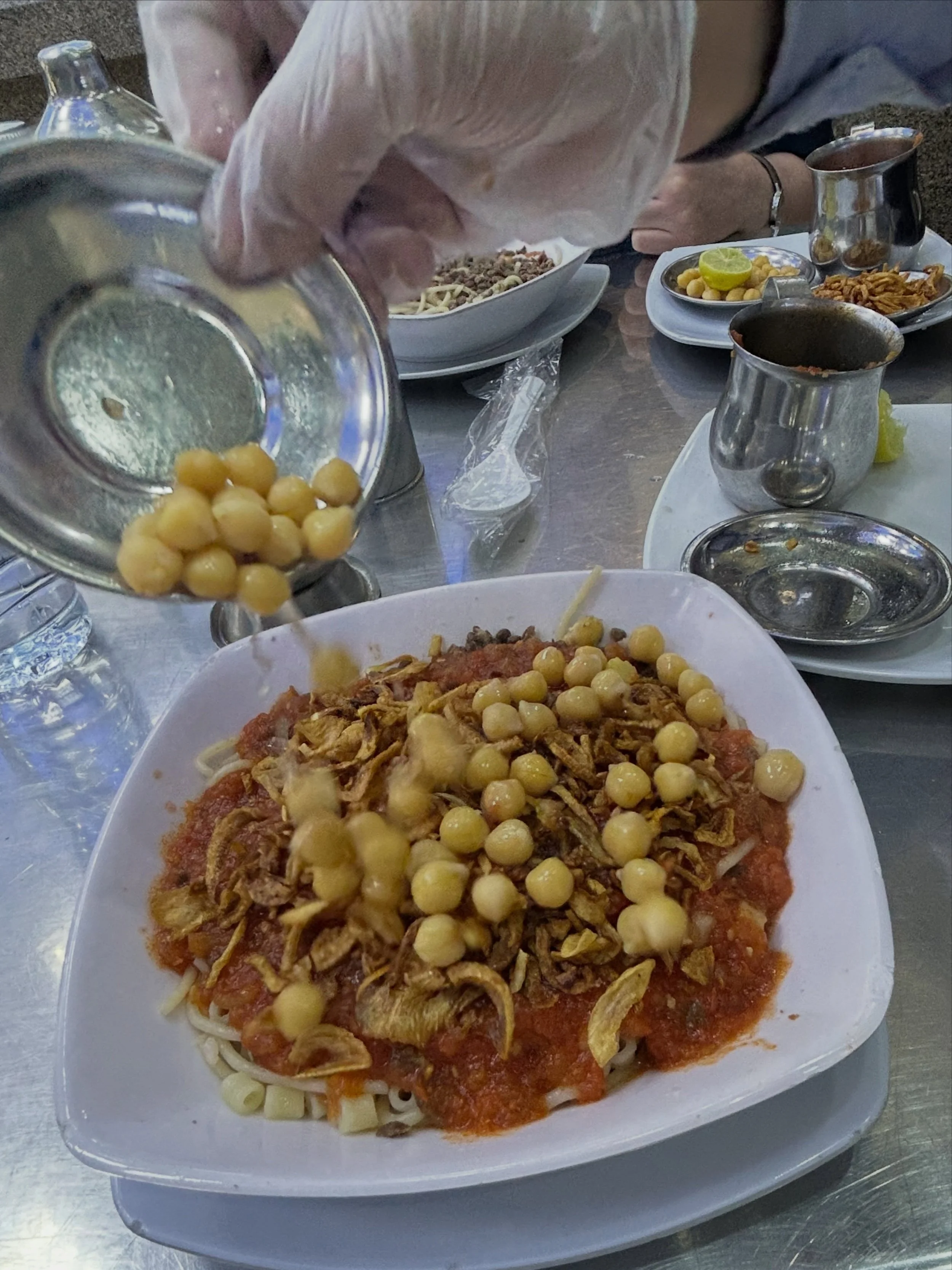 Whoever thought of combining macaroni, fried rice, vermicelli, brown lentils + topping the heap with tomato sauce, crispy fried onions, chickpeas, garlic vinegar + hot sauce was a carb-loving genius. We&rsquo;ve heard different origin stories, but wh