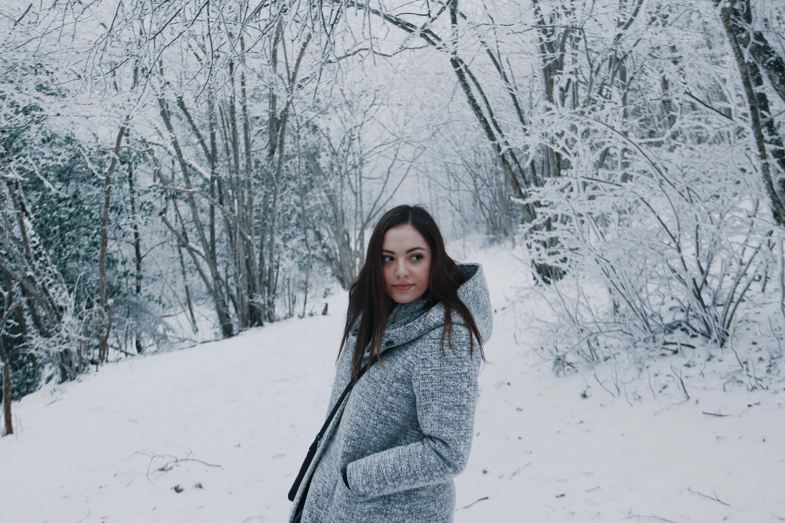 Une femme en manteau gris dans une forêt enneigée, arbres recouverts de neige.