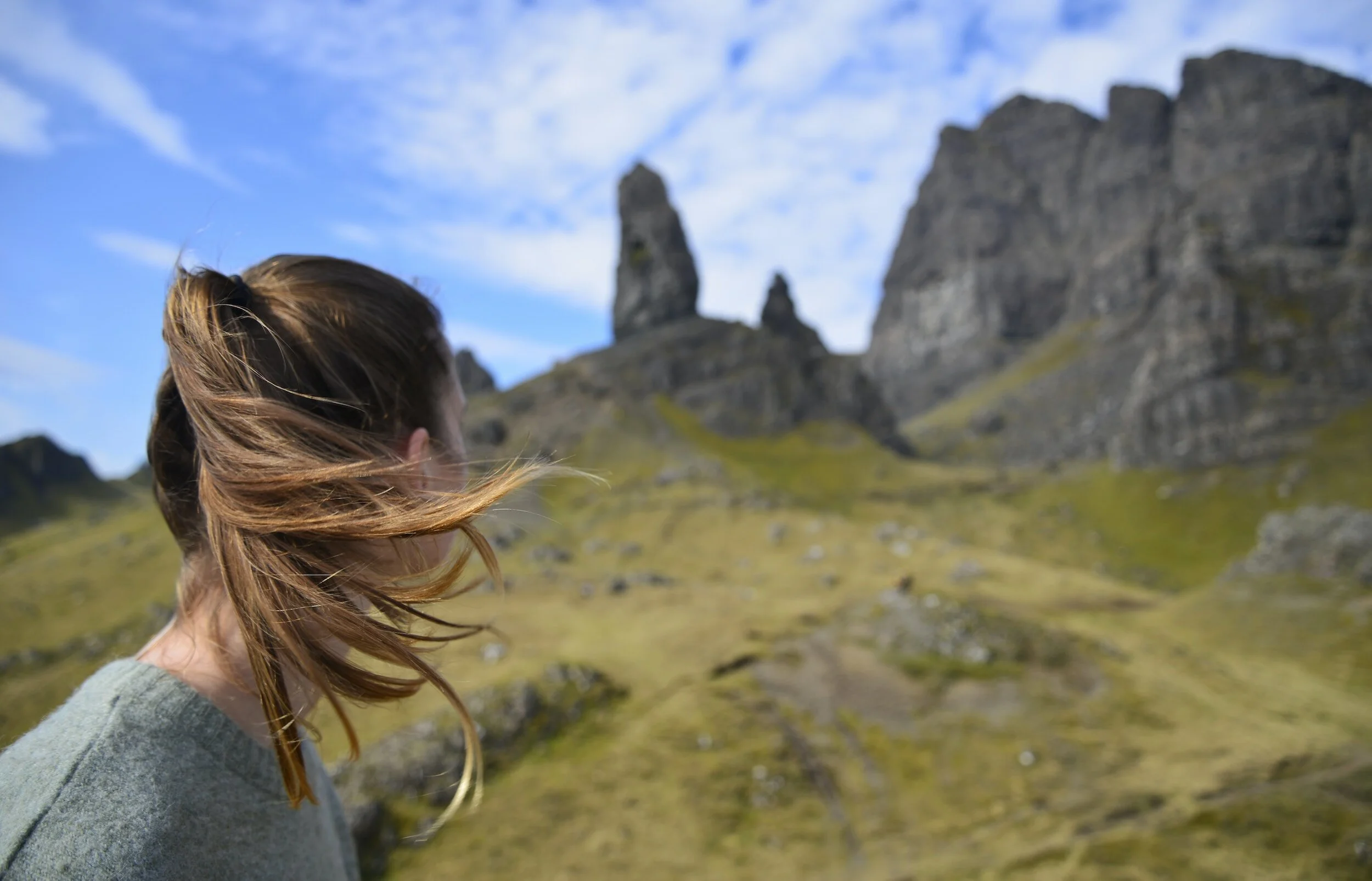 Une personne avec des cheveux bruns se tient devant un paysage montagneux, avec des pics rocheux et un ciel nuageux en arrière-plan.