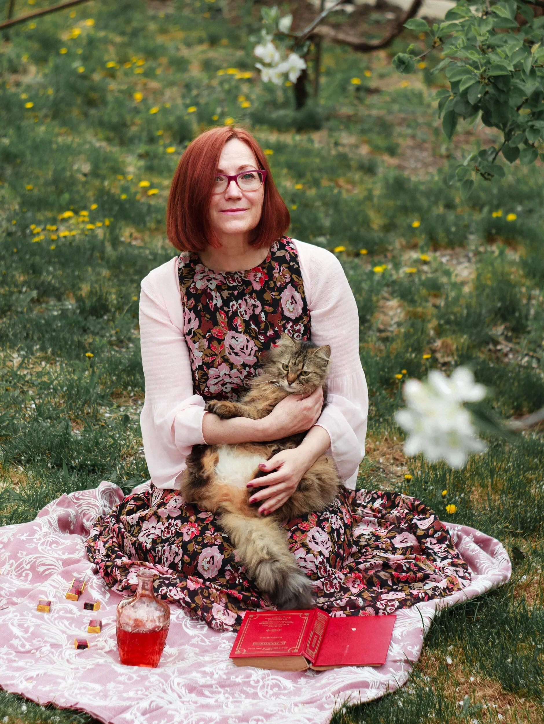 Une femme aux cheveux roux et portant des lunettes, assise dans un jardin sur une couverture rose à motifs floraux. Elle tient un chat à poil long sur ses genoux. Autour d'elle, il y a un livre rouge ouvert et une carafe contenant une boisson rouge. On aperçoit des fleurs et de l'herbe avec des pissenlits dans le fond.