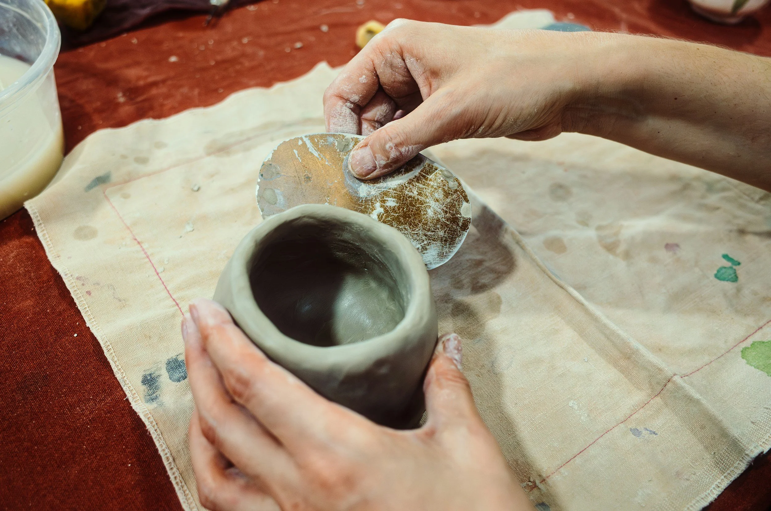 Main tenant sculptant un petit bol en argile avec un outil de sculpture, sur un tissu peint à la main, au-dessus d'une table en bois.