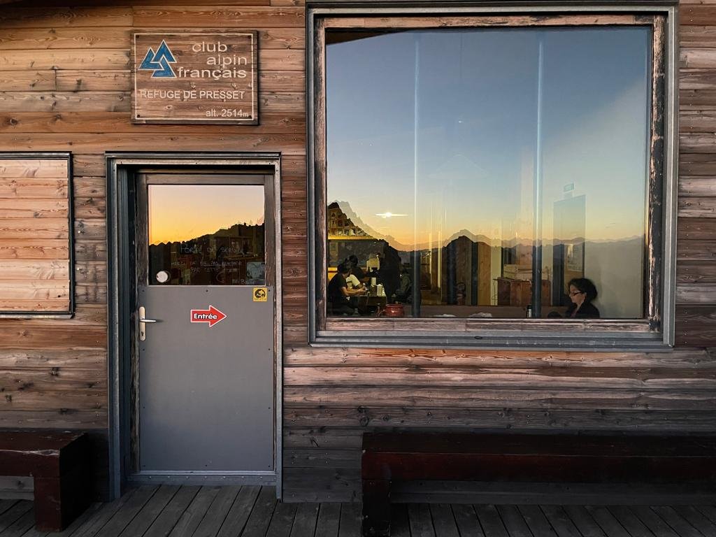 Refuge de montagne en bois avec une fenêtre reflétant le coucher de soleil et l'enseigne "Club Alpin Français, Refuge de Presset, alt. 2514 m". Une porte avec une flèche et "Entrée" est visible.