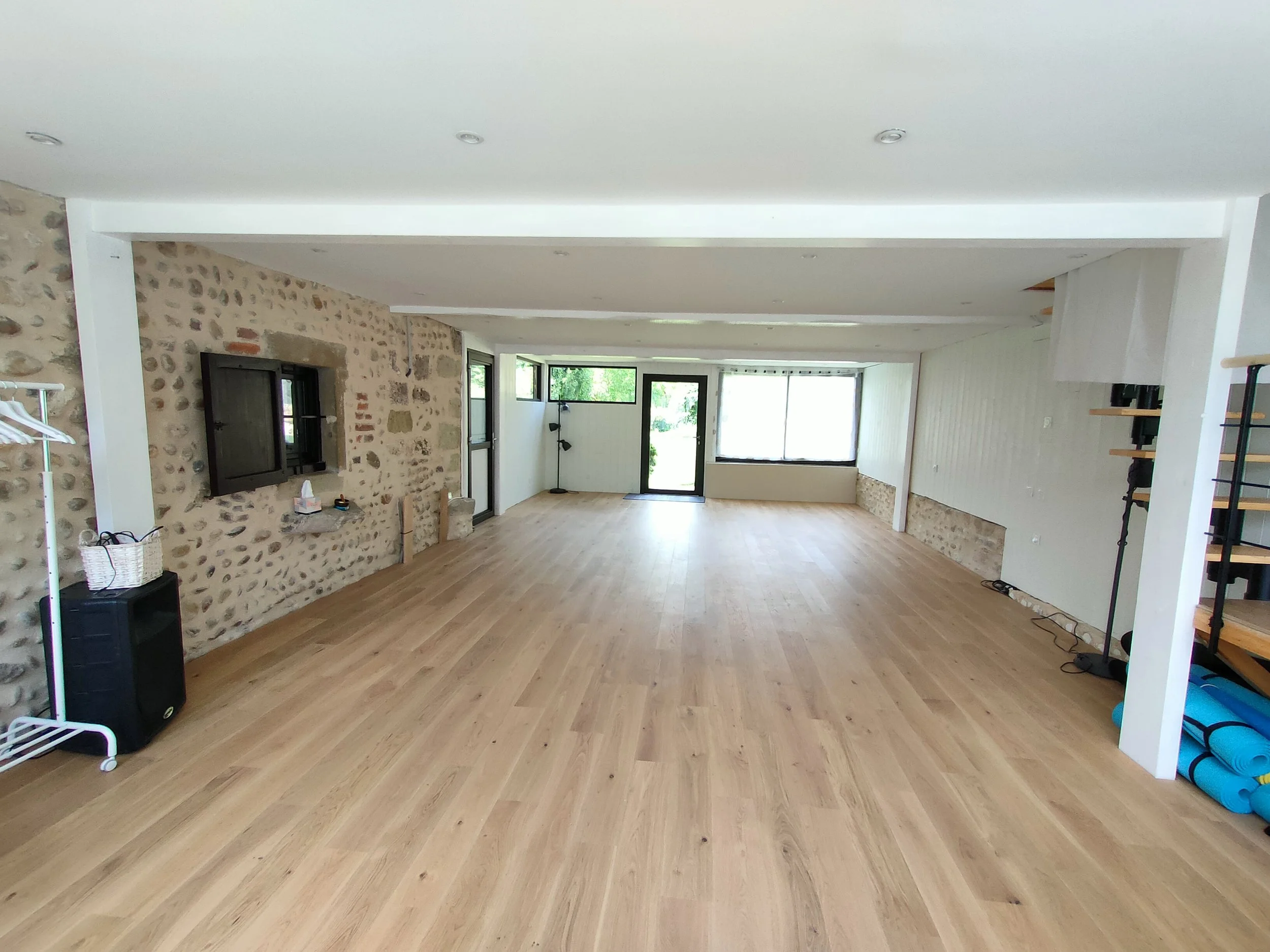 Grand espace intérieur avec sol en bois clair, mur en pierre et murs blancs, grandes fenêtres laissant entrer la lumière naturelle, un escalier avec rampes en métal et bois, et quelques meubles ainsi que tapis de yoga enroulés