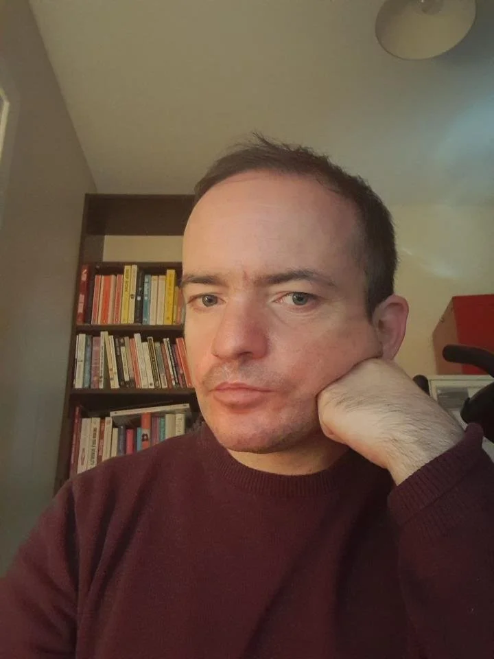A man with short dark hair resting his head on his hand in a room with a bookshelf filled with books in the background.