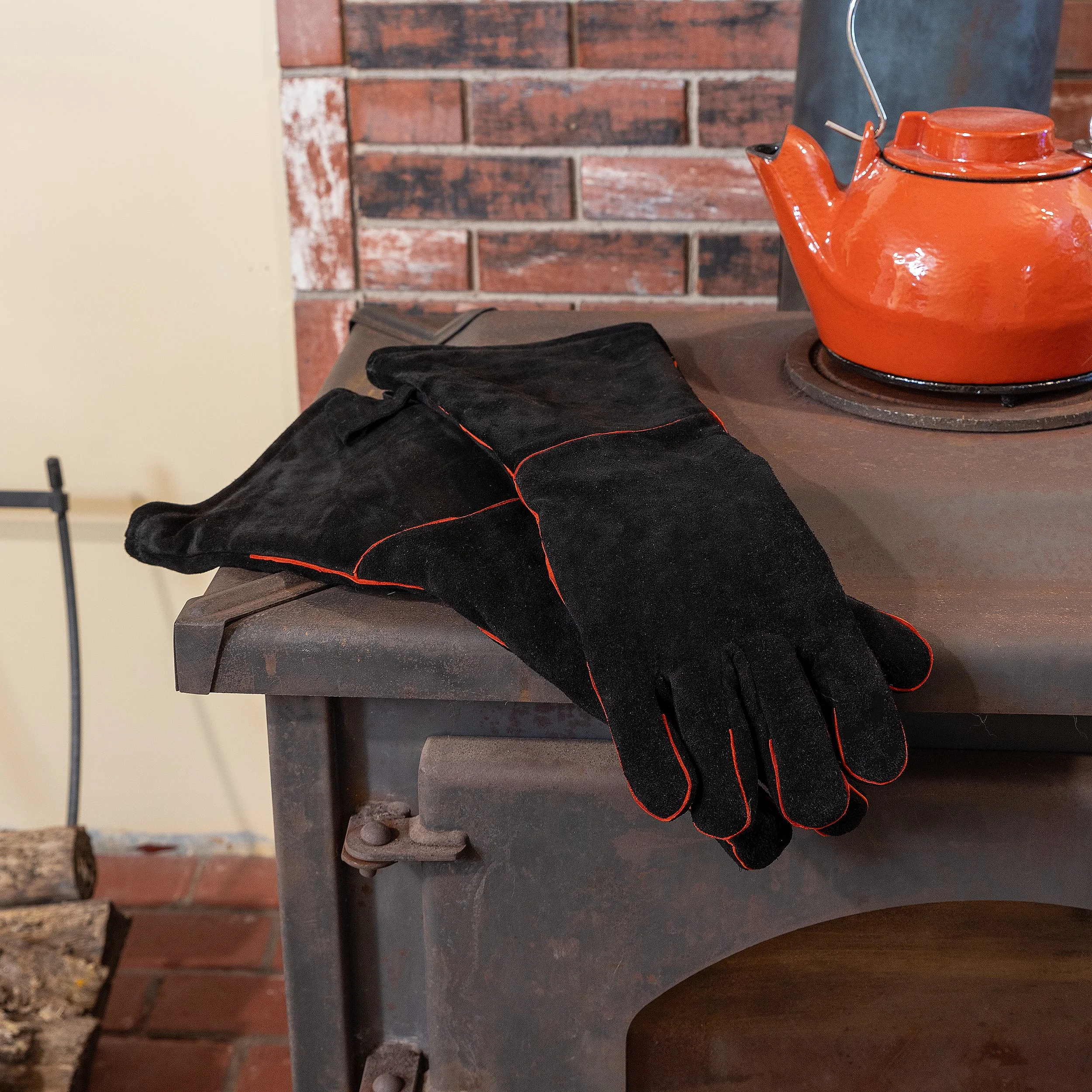 Black heat-resistant gloves with red piping resting on a metal stove near a red steemer in a kitchen with a brick wall background.