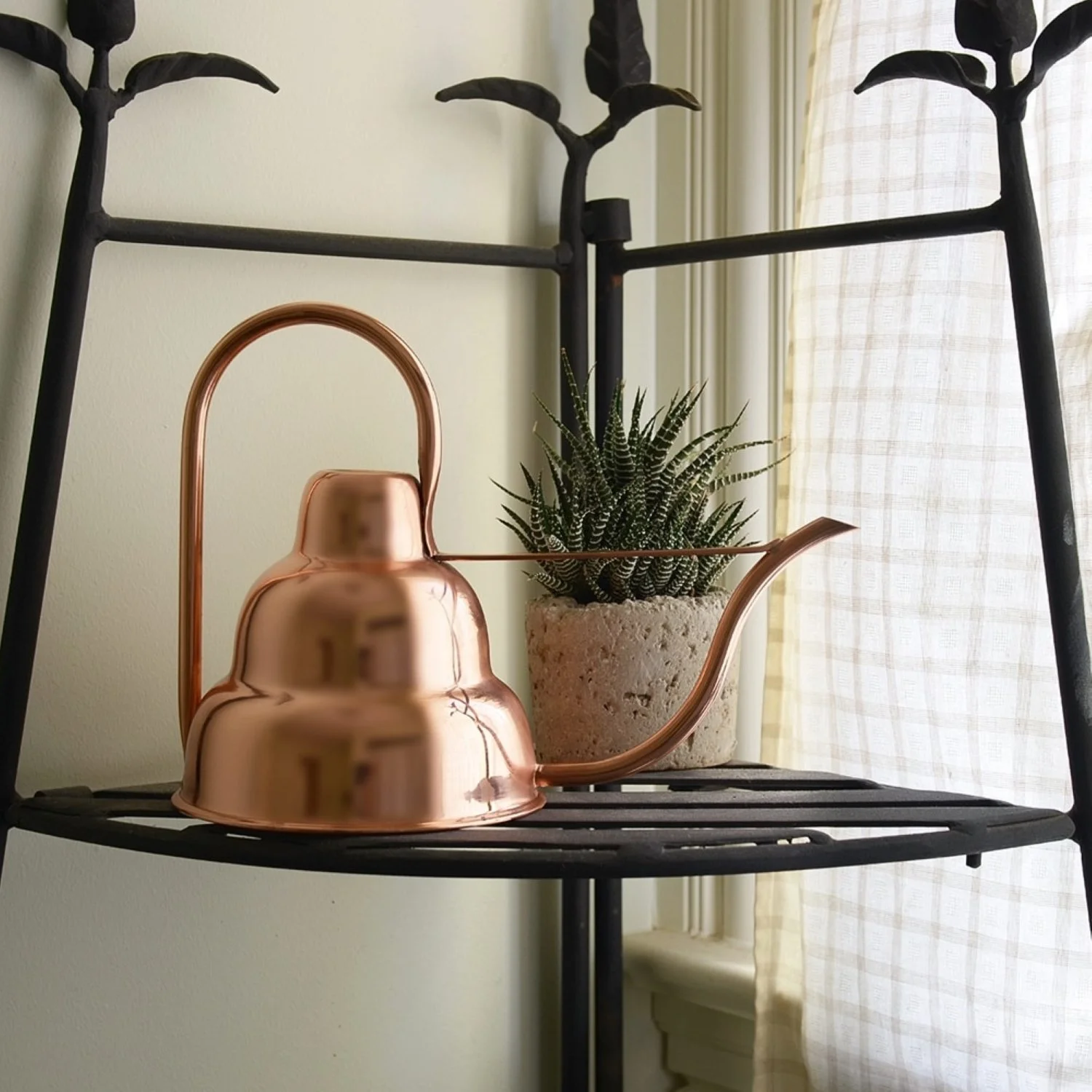 Teared art deco style solid spun copper watering can resting on a bathroom shelf next to a window