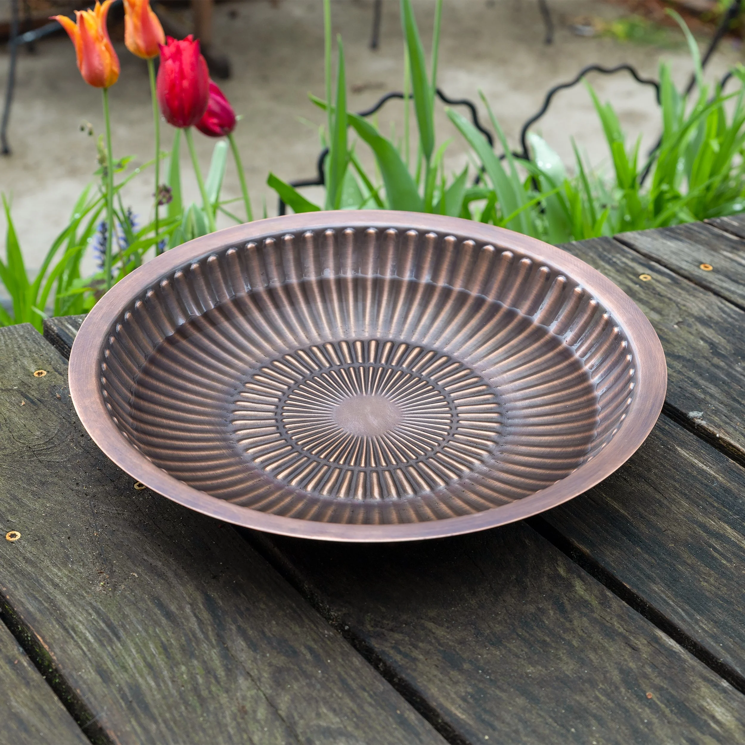 A decorative metal plate with a radial pattern, placed on a weathered wooden surface outdoors surrounded by green plants and colorful tulips in the background.