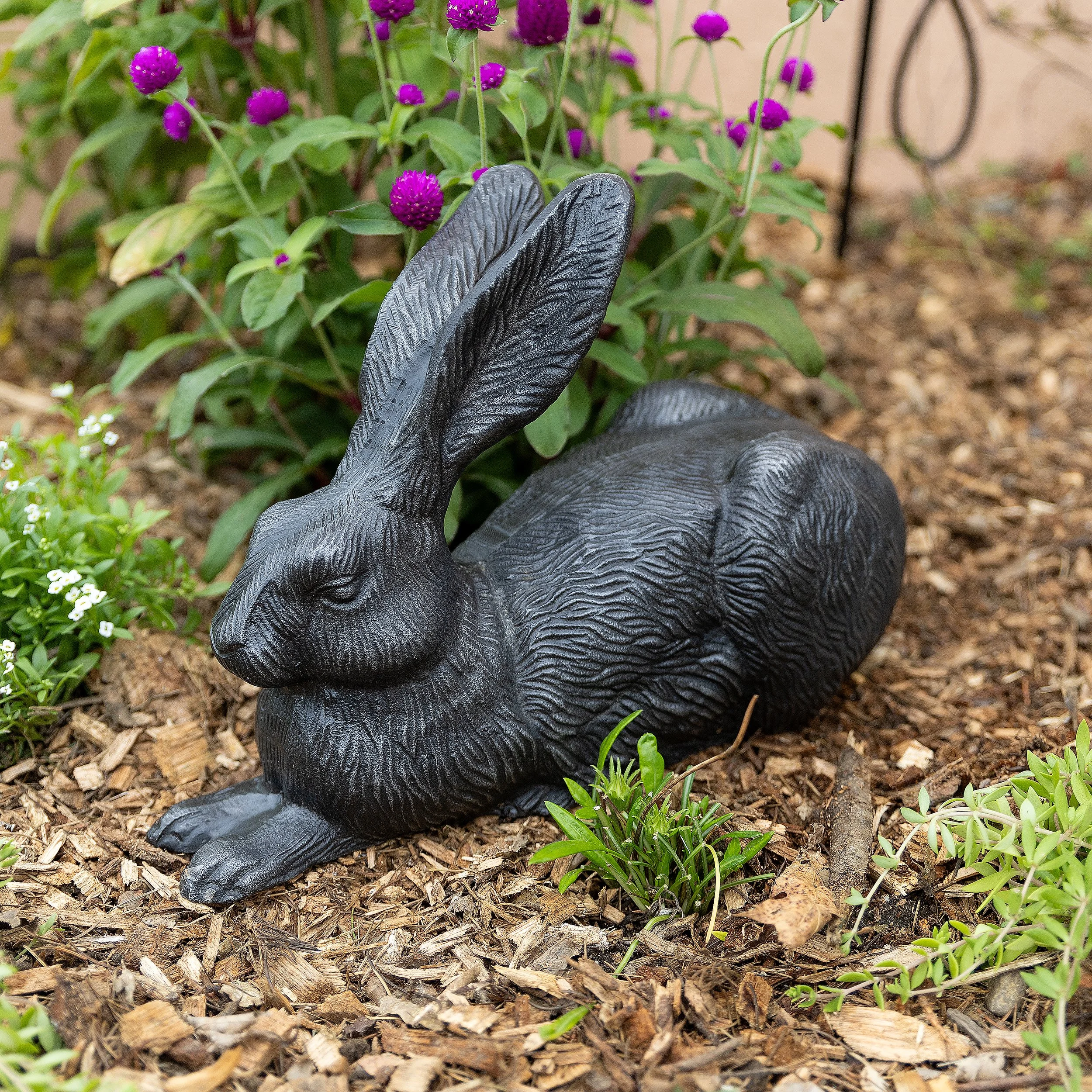 Black rabbit ornament placed on mulch garden bed with purple and white flowers in the background.