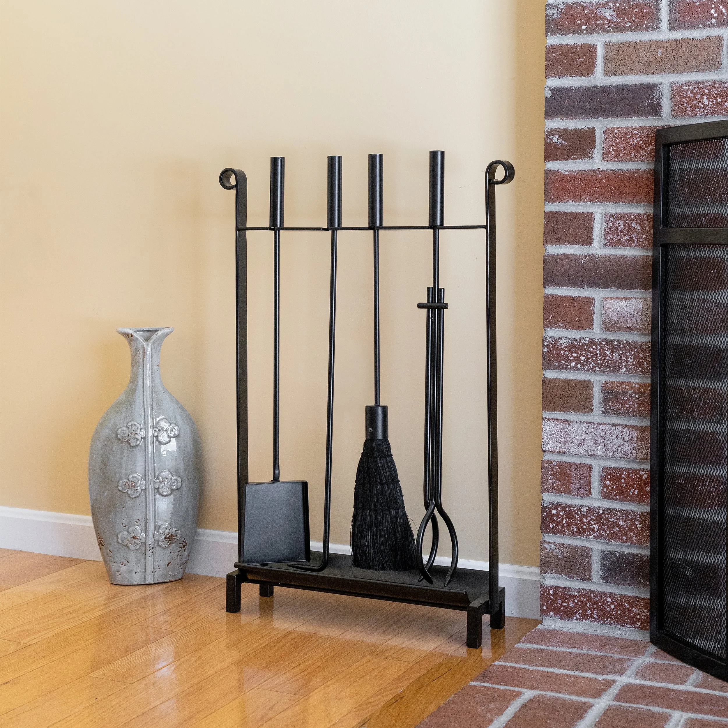 Fireplace tools set including a brush, poker, tongs, and shovel next to a decorative vase on the wooden floor.