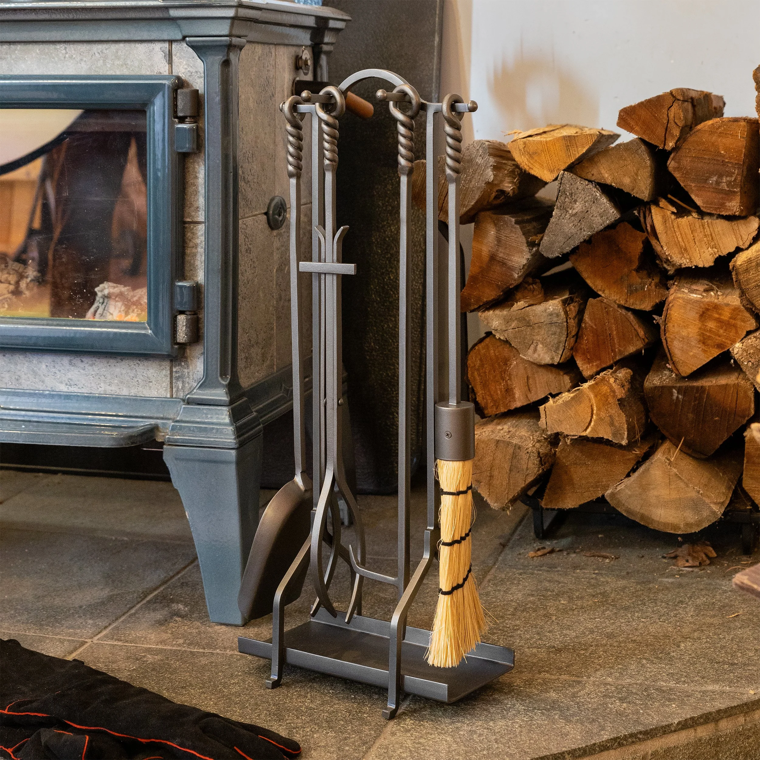 A set of metal fireplace tools, including a poker, broom, and tongs, leaning against a dark fireplace with a wood-burning stove. A stack of chopped firewood is stored nearby on the right side.
