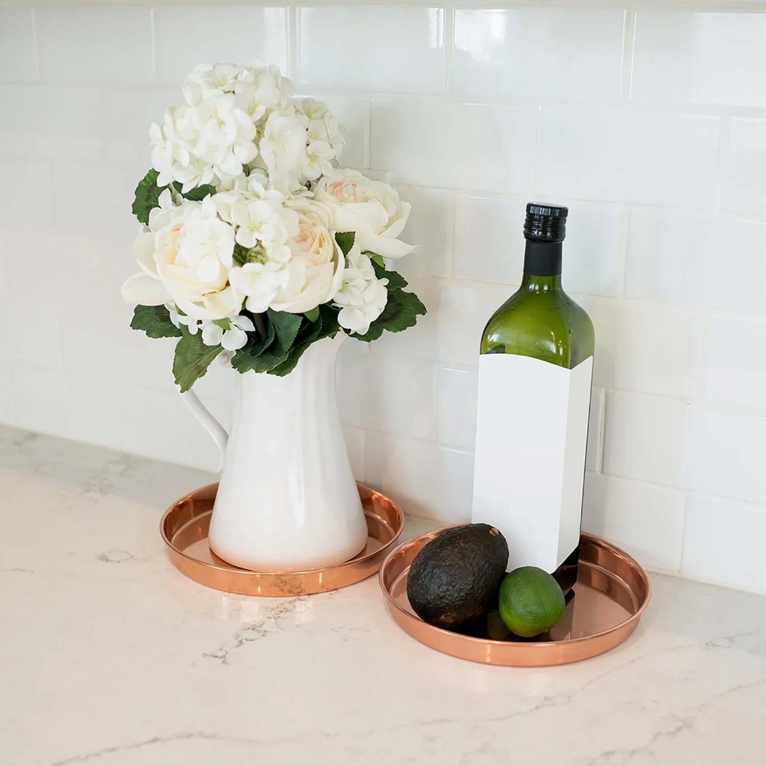 Trays pictured on a kitchen counter top. Olive oil bottle and avocados on one tray. the second tray has a pitcher with flowers in it.