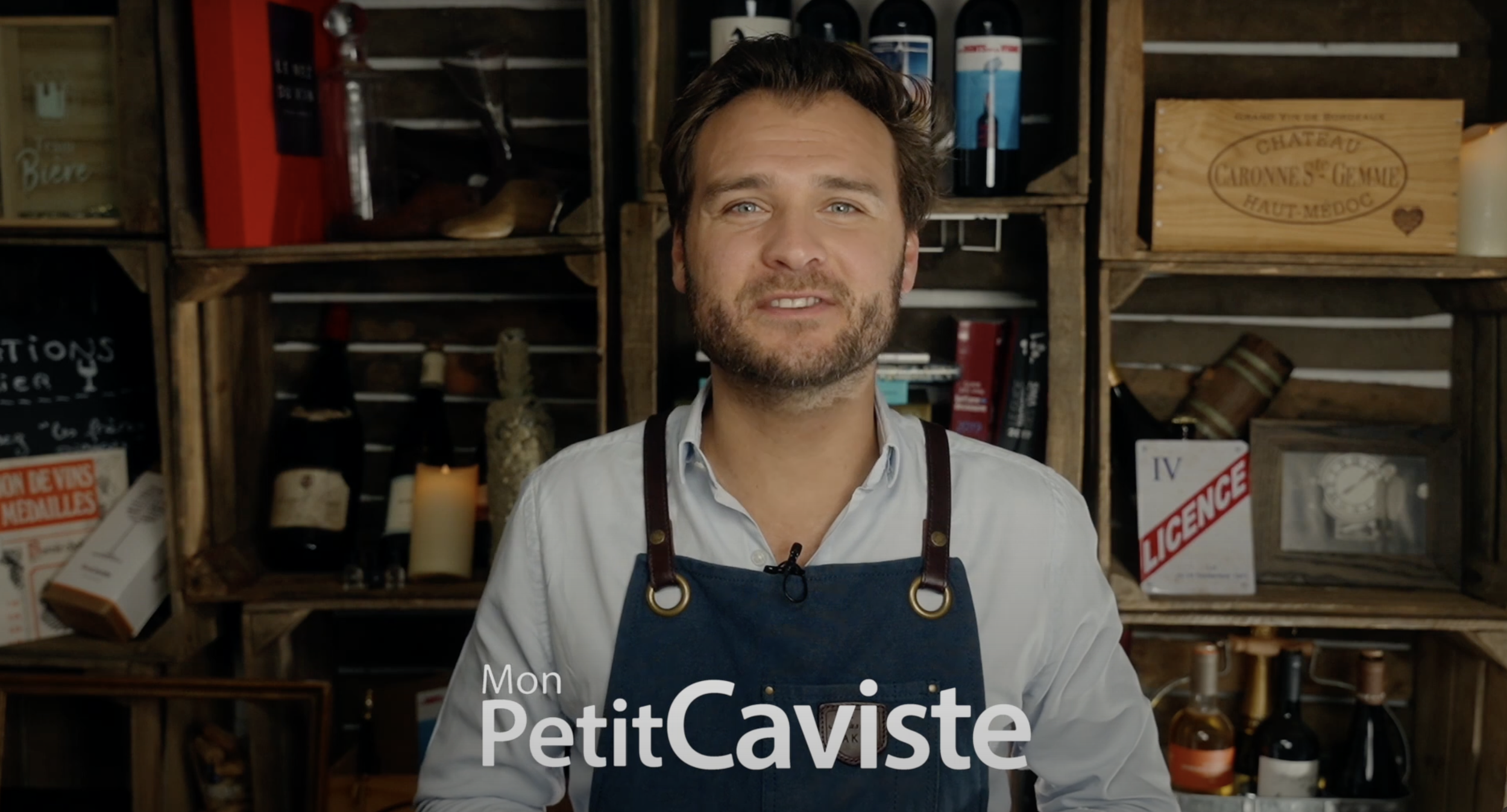 Un homme souriant portant un tablier en cuir dans une cave à vin ou un espace de dégustation de vin, avec des bouteilles et des caisses en bois en arrière-plan, écrit en français : "Mon PetitCaviste".