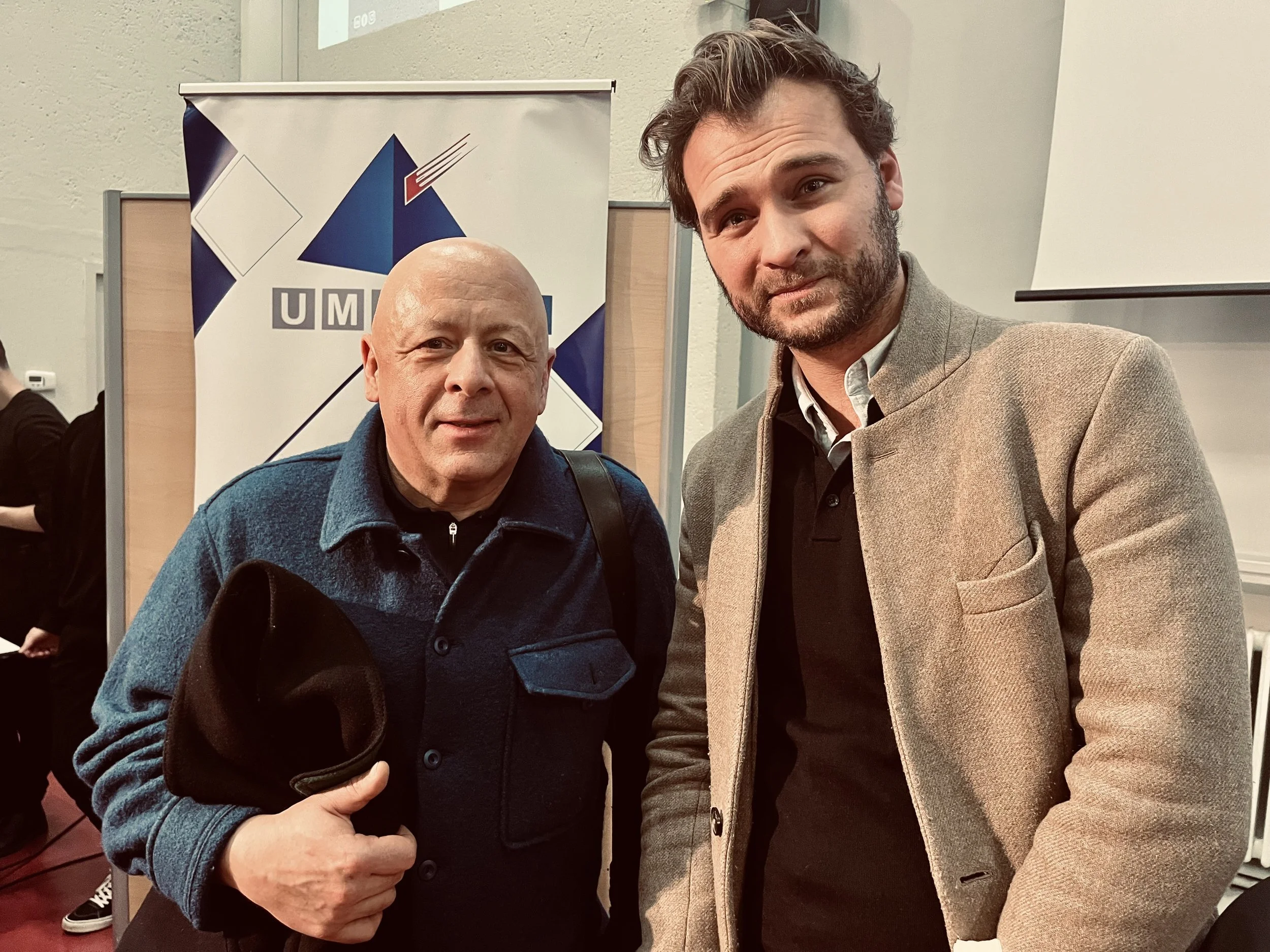 Deux hommes posent pour une photo, l'un porte un manteau beige et l'autre un sweat bleu foncé, à l'arrière-plan un logo UMS et un tableau blanc.