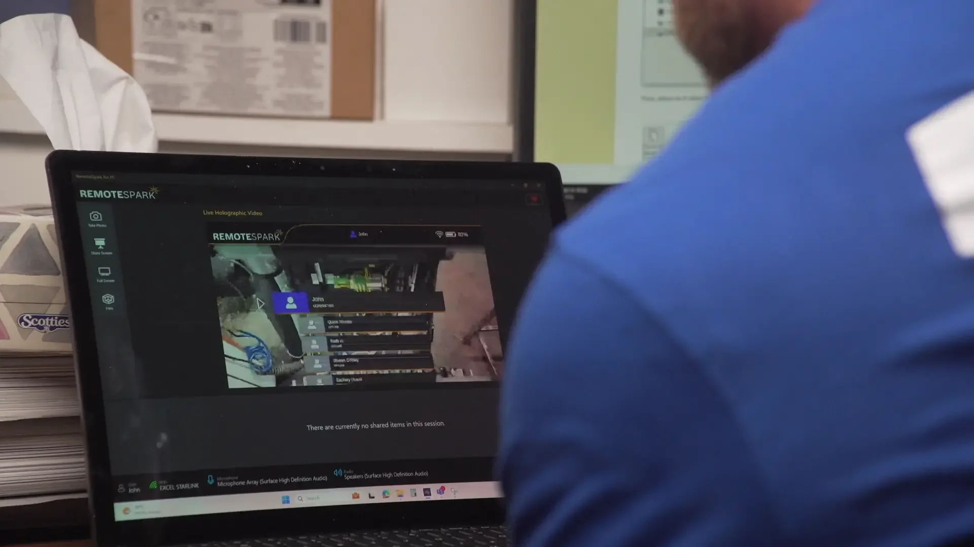 A person in a blue shirt using a laptop computer with a software interface labeled 'RemoteSpark' on the screen, showing a live holographic video interface and a list of connected users.