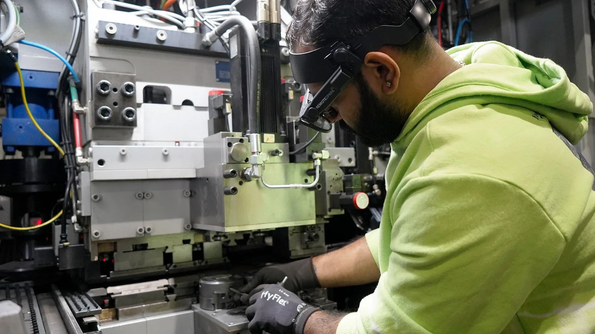 A man wearing Augmented Reality glasses and a green hoodie working on a piece of electronic or mechanical equipment with various wires and components.