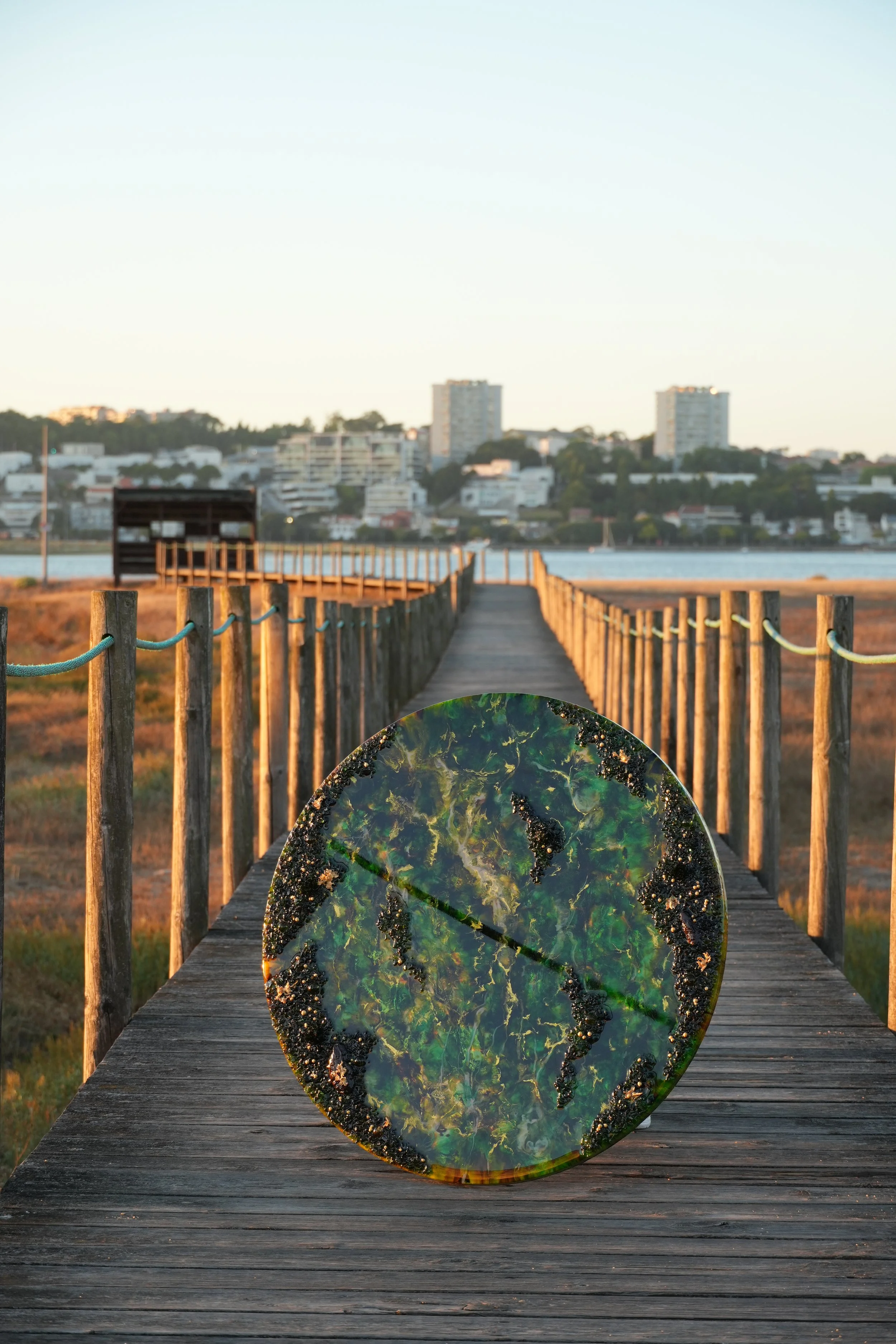 Imagem de uma ponte de madeira com uma peça de vidro de arte com cores verdes, pretas e douradas, na frente, ao pôr do sol com uma cidade ao fundo.